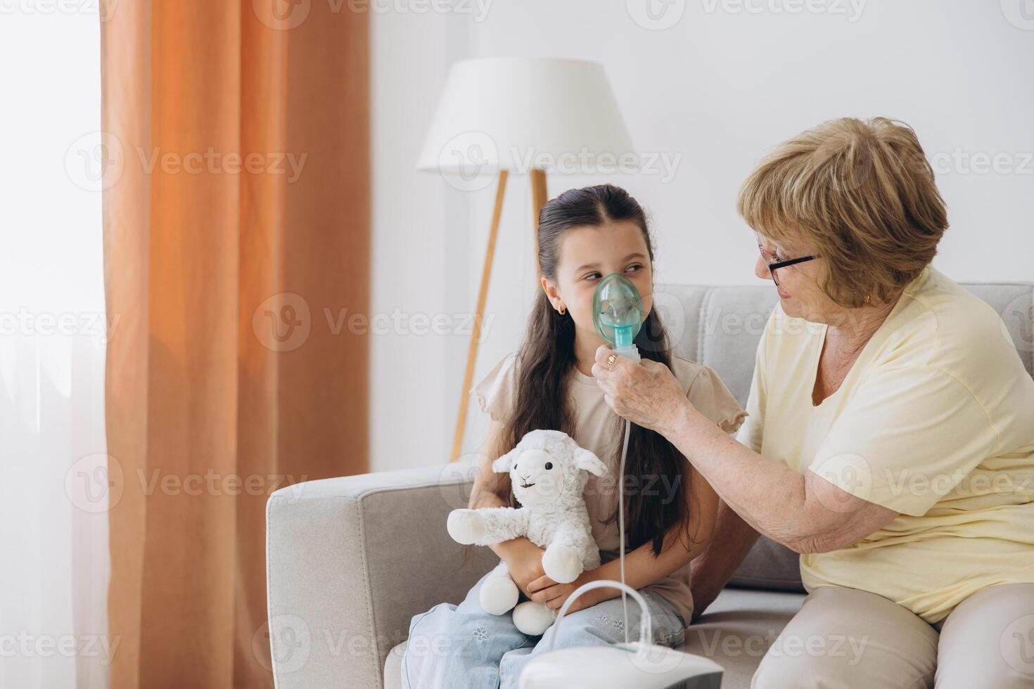 Little girl making inhalation with nebulizer with granny. Woman makes inhalation to a sick child while embracing her. Grandmother helping granddaughter with cold and flu to inahale nebuliser aerosol. photo