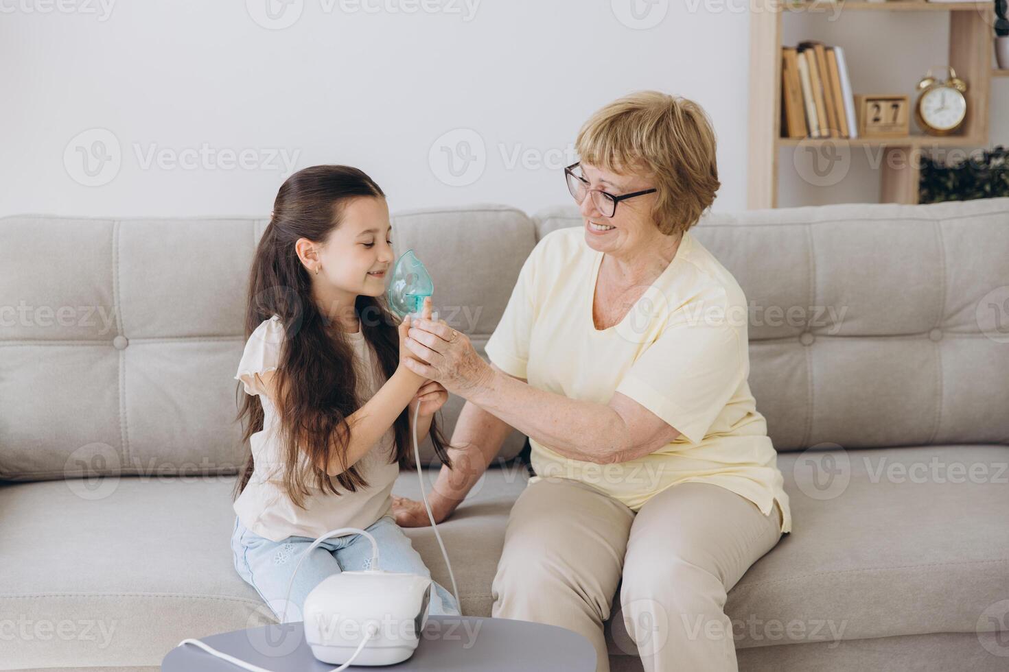 Little girl making inhalation with nebulizer with granny. Woman makes inhalation to a sick child while embracing her. Grandmother helping granddaughter with cold and flu to inahale nebuliser aerosol. photo