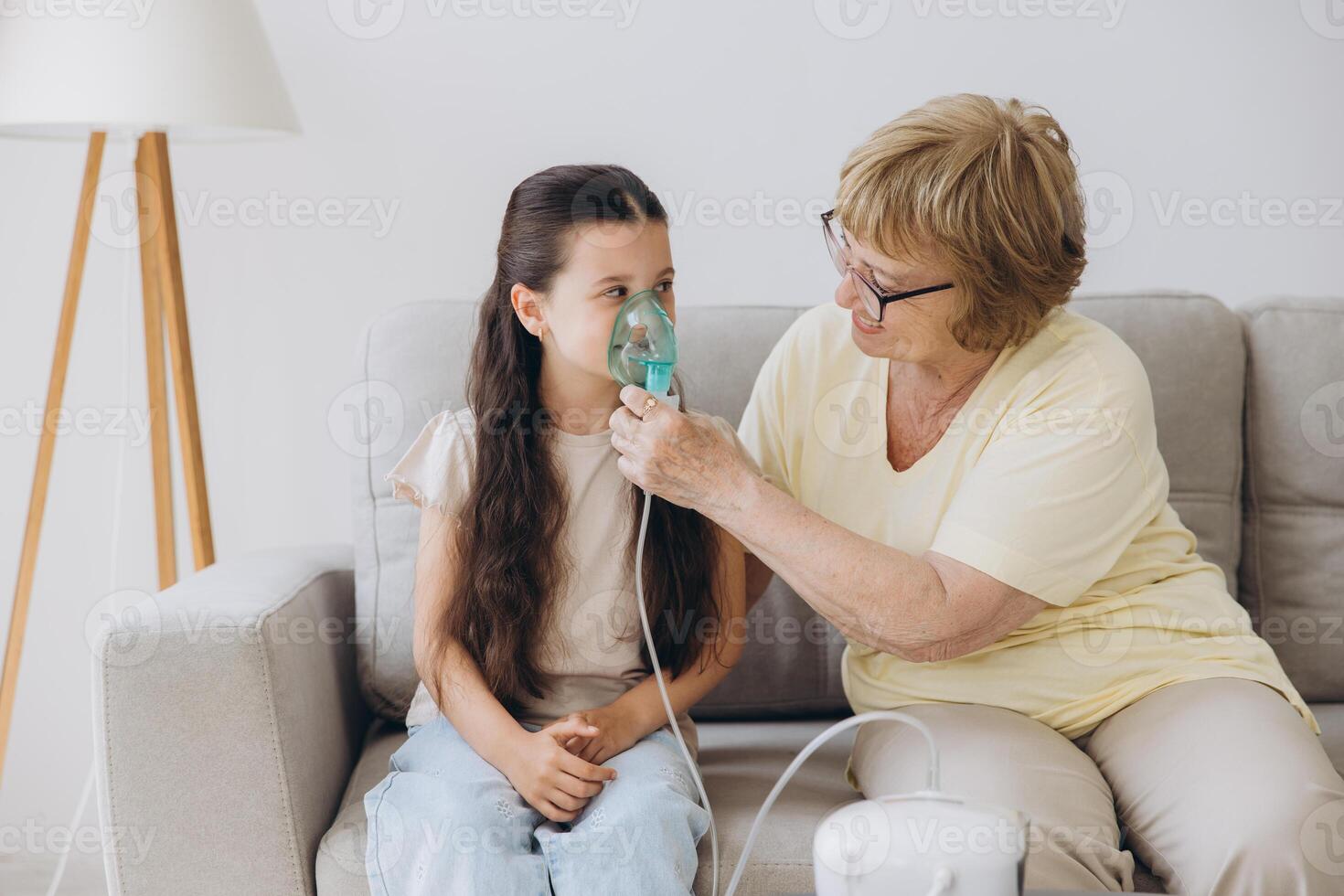 Little girl making inhalation with nebulizer with granny. Woman makes inhalation to a sick child while embracing her. Grandmother helping granddaughter with cold and flu to inahale nebuliser aerosol. photo