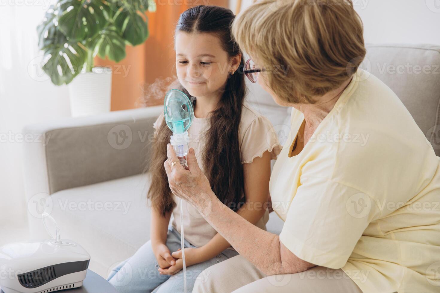 Little girl making inhalation with nebulizer with granny. Woman makes inhalation to a sick child while embracing her. Grandmother helping granddaughter with cold and flu to inahale nebuliser aerosol. photo
