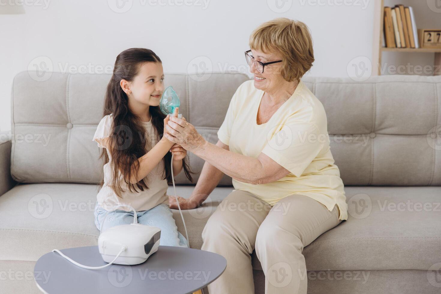 Little girl making inhalation with nebulizer with granny. Woman makes inhalation to a sick child while embracing her. Grandmother helping granddaughter with cold and flu to inahale nebuliser aerosol. photo