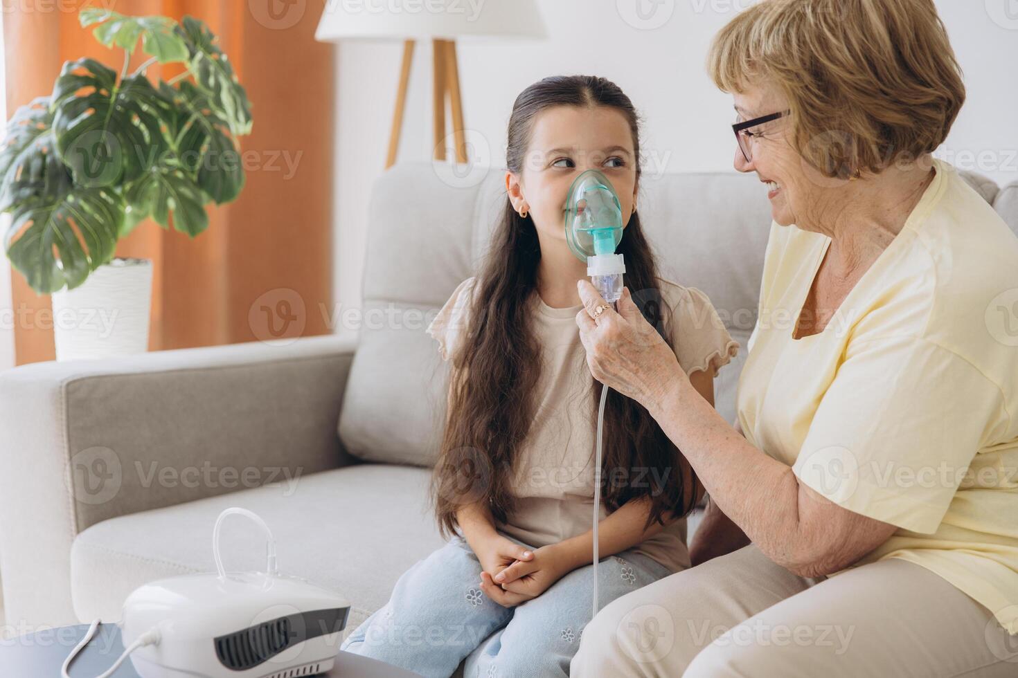 Little girl making inhalation with nebulizer with granny. Woman makes inhalation to a sick child while embracing her. Grandmother helping granddaughter with cold and flu to inahale nebuliser aerosol. photo
