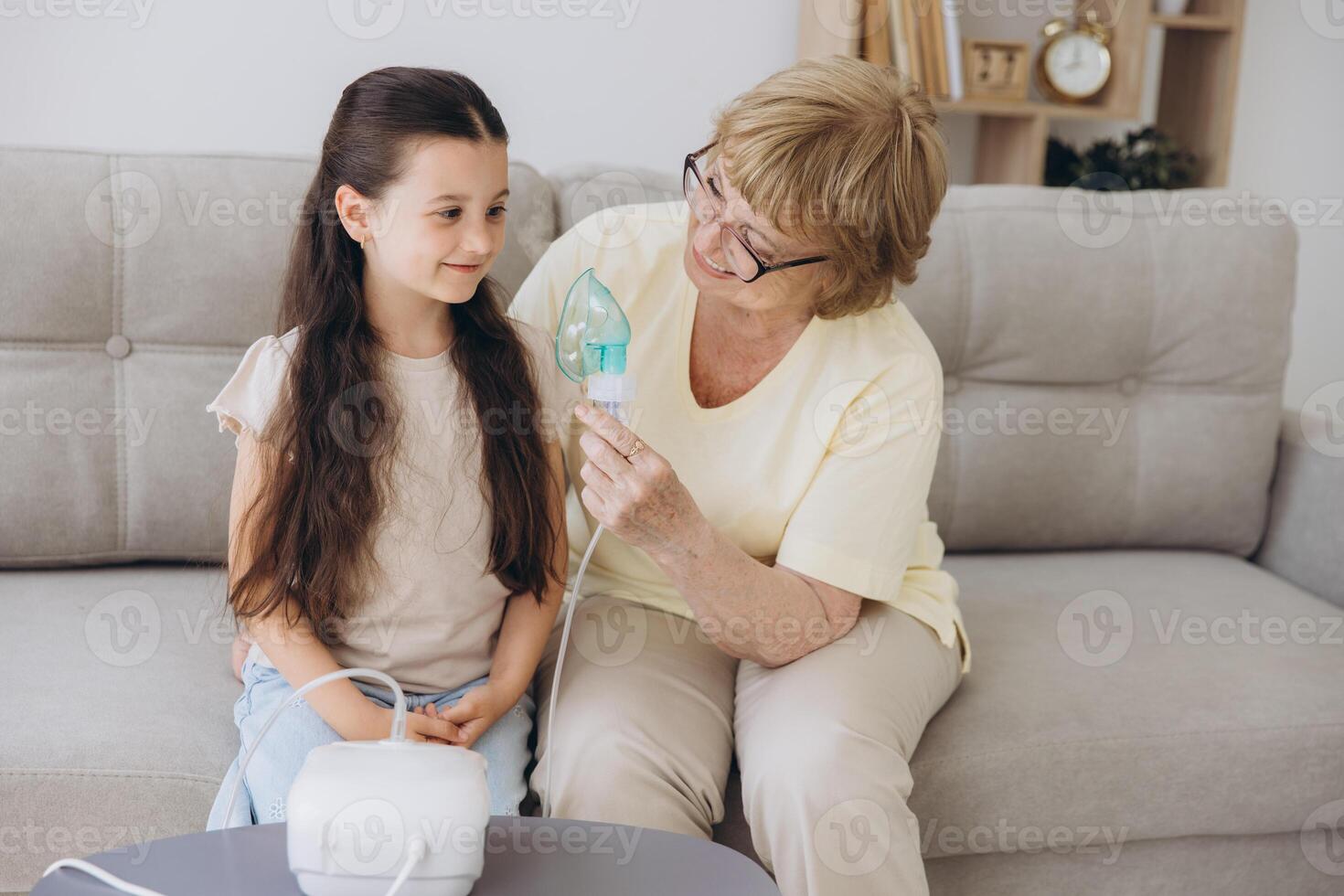 Little girl making inhalation with nebulizer with granny. Woman makes inhalation to a sick child while embracing her. Grandmother helping granddaughter with cold and flu to inahale nebuliser aerosol. photo