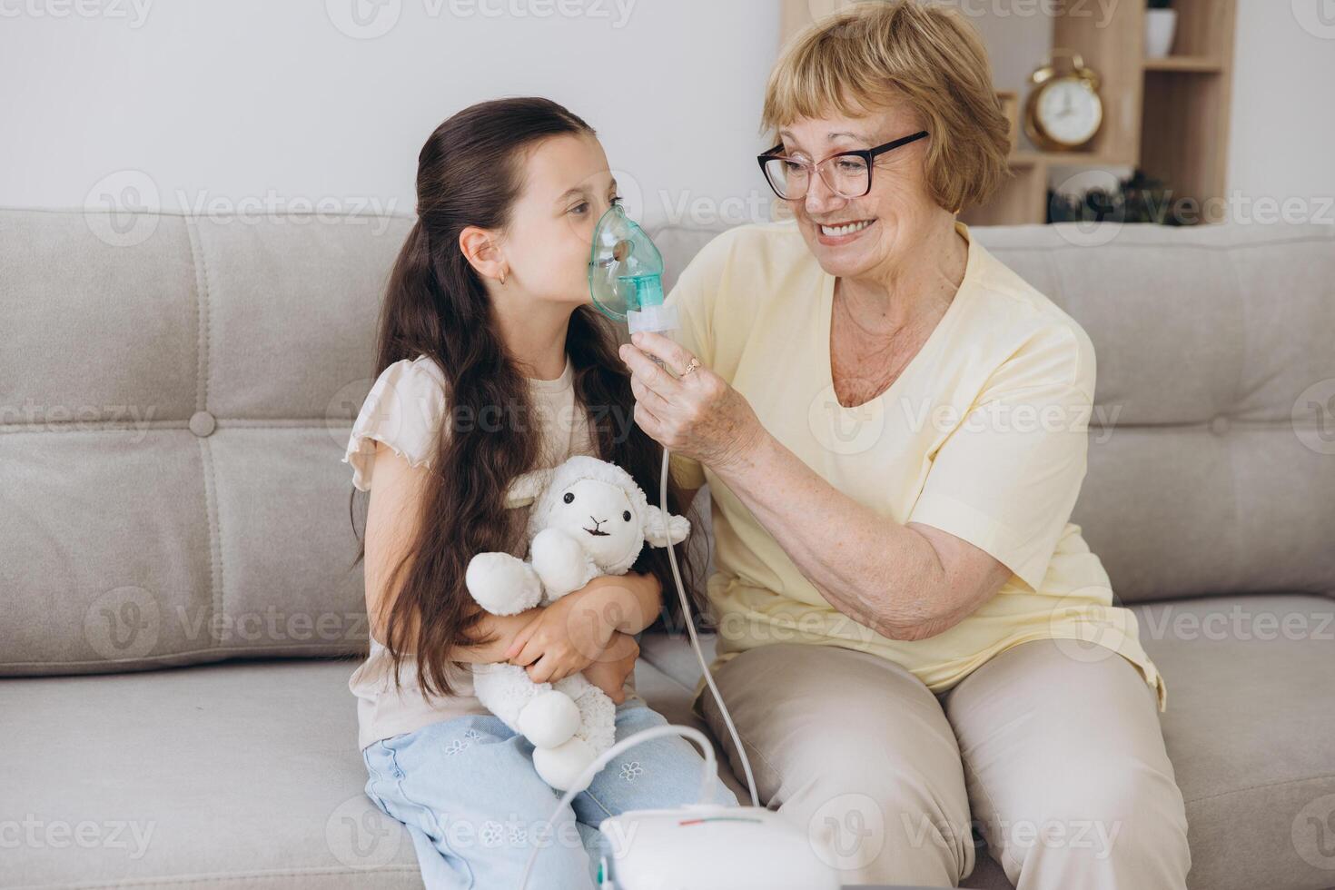 Little girl making inhalation with nebulizer with granny. Woman makes inhalation to a sick child while embracing her. Grandmother helping granddaughter with cold and flu to inahale nebuliser aerosol. photo