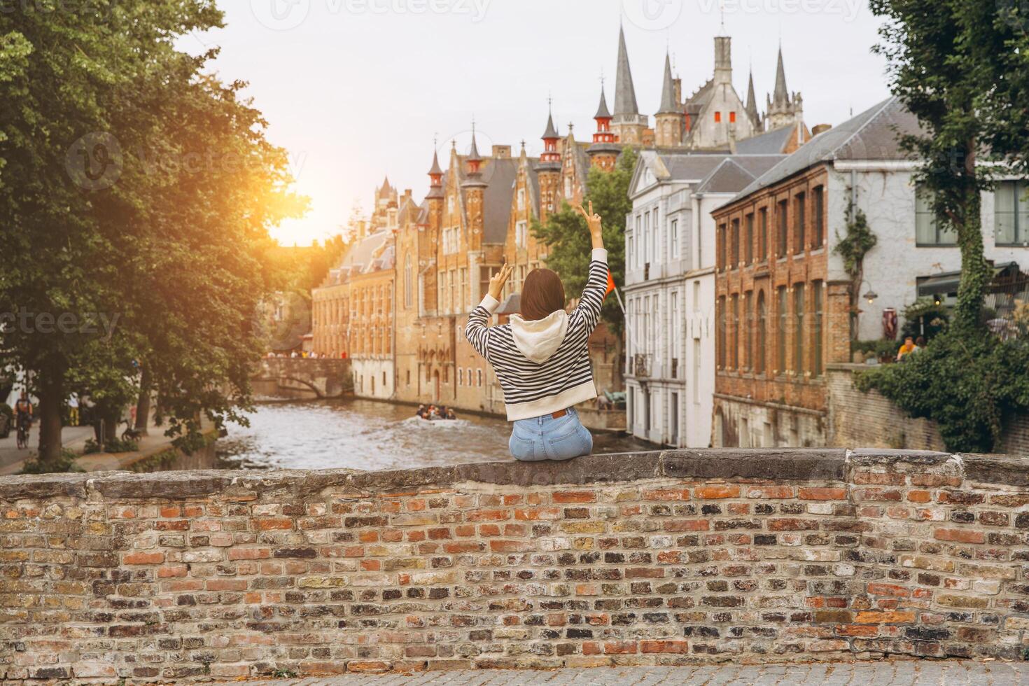 Girl tourist sits on the bridge overlooking the canal and admires the view of the picturesque and historical city of Bruges at sunset, Belgium photo