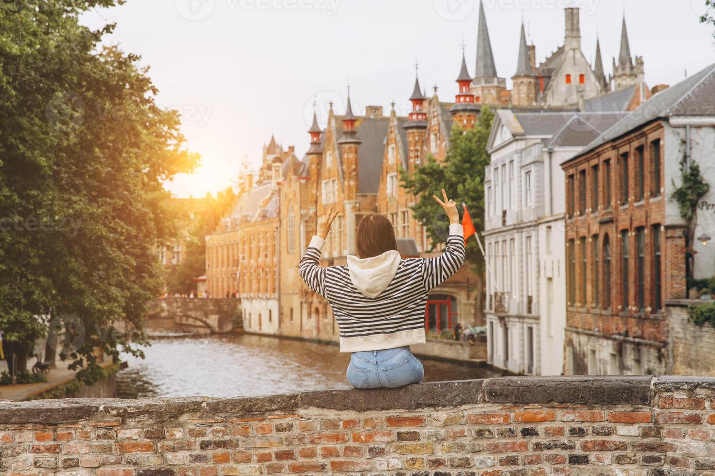 Girl tourist sits on the bridge overlooking the canal and admires the view of the picturesque and historical city of Bruges at sunset, Belgium photo