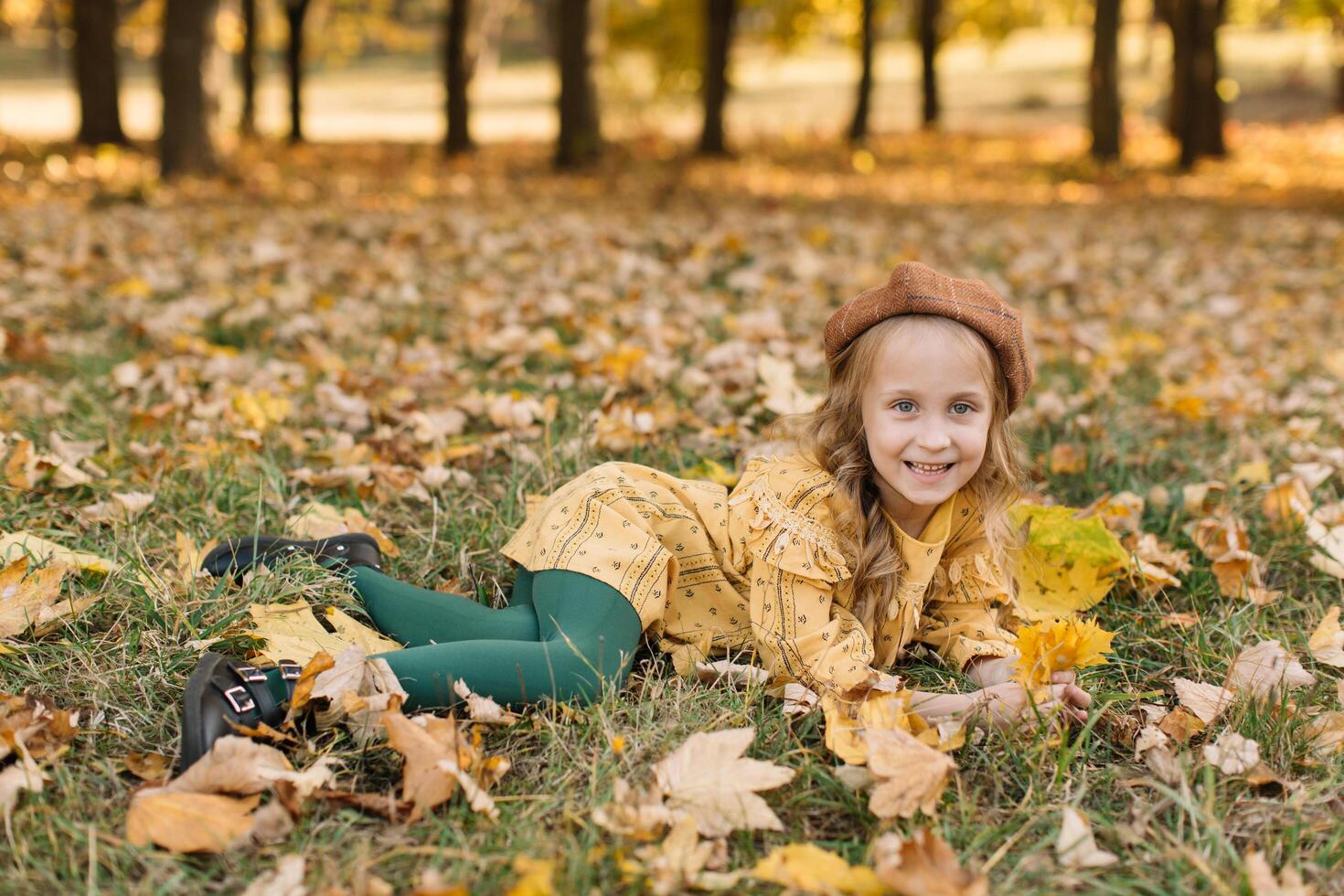 Portrait of a beautiful little blonde girl in a yellow dress in an autumn park lying in the middle of maple leaves. photo