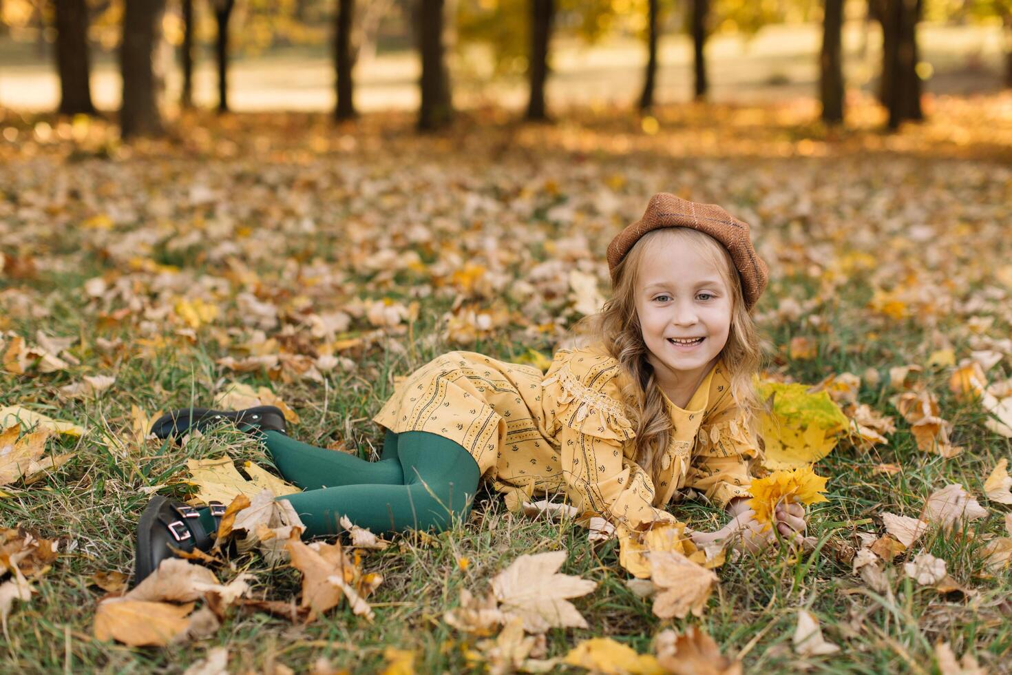 Portrait of a beautiful little blonde girl in a yellow dress in an autumn park lying in the middle of maple leaves. photo