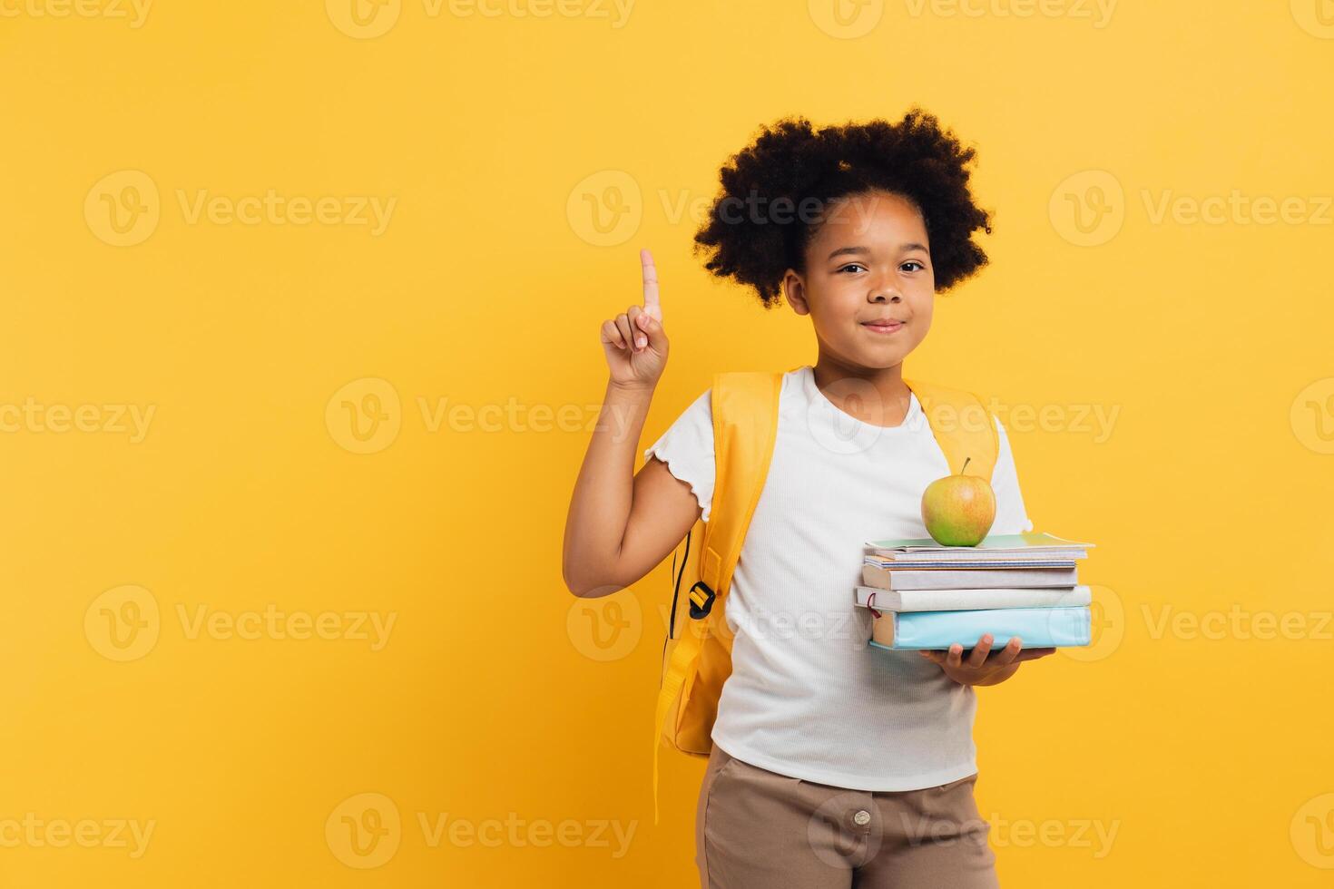 African american schoolgirl holding notebooks and books and showing something on yellow background, copy space. photo