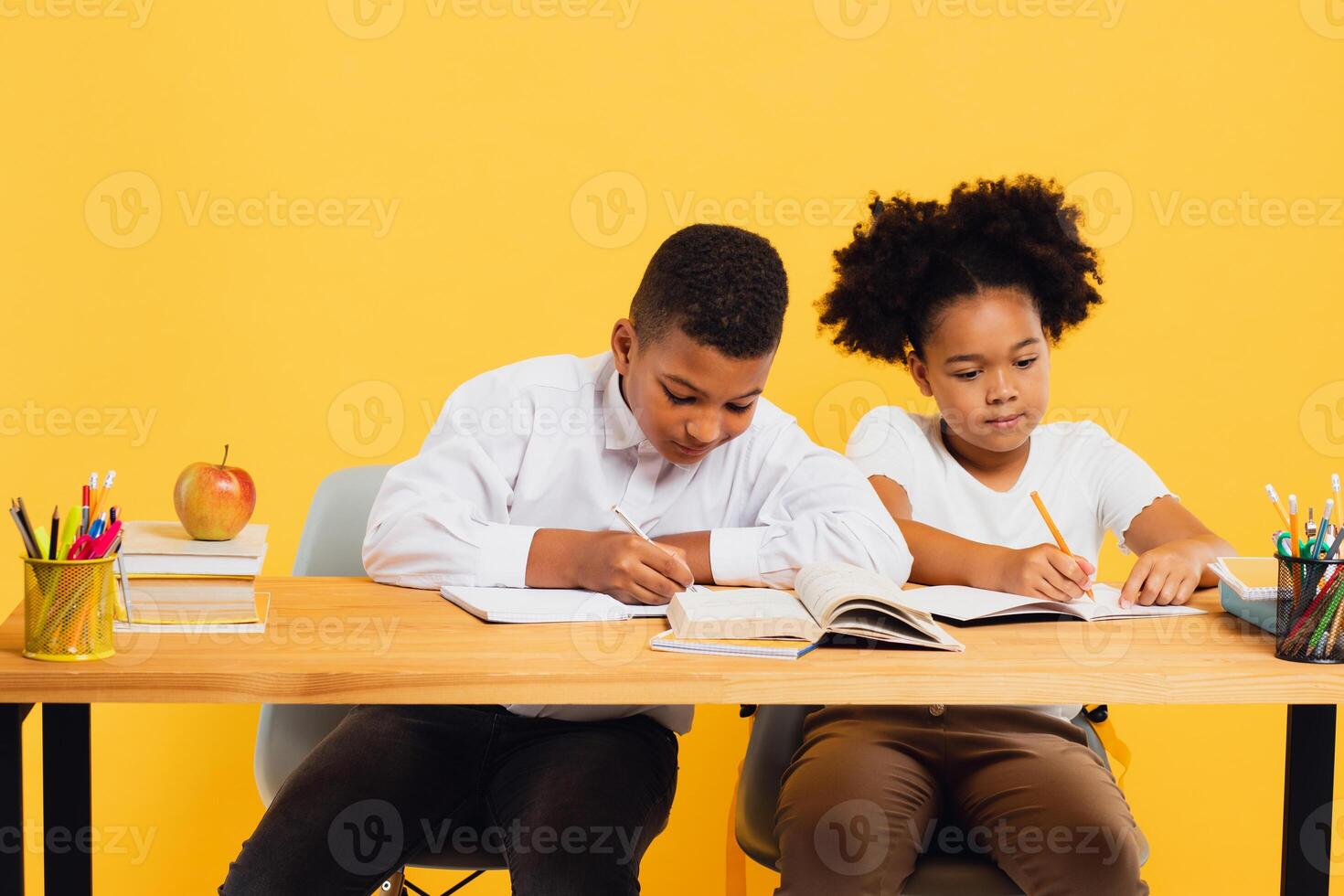 Happy african american schoolgirl and mixed race schoolboy sitting together at desk and studying on yellow background. Back to school concept. photo