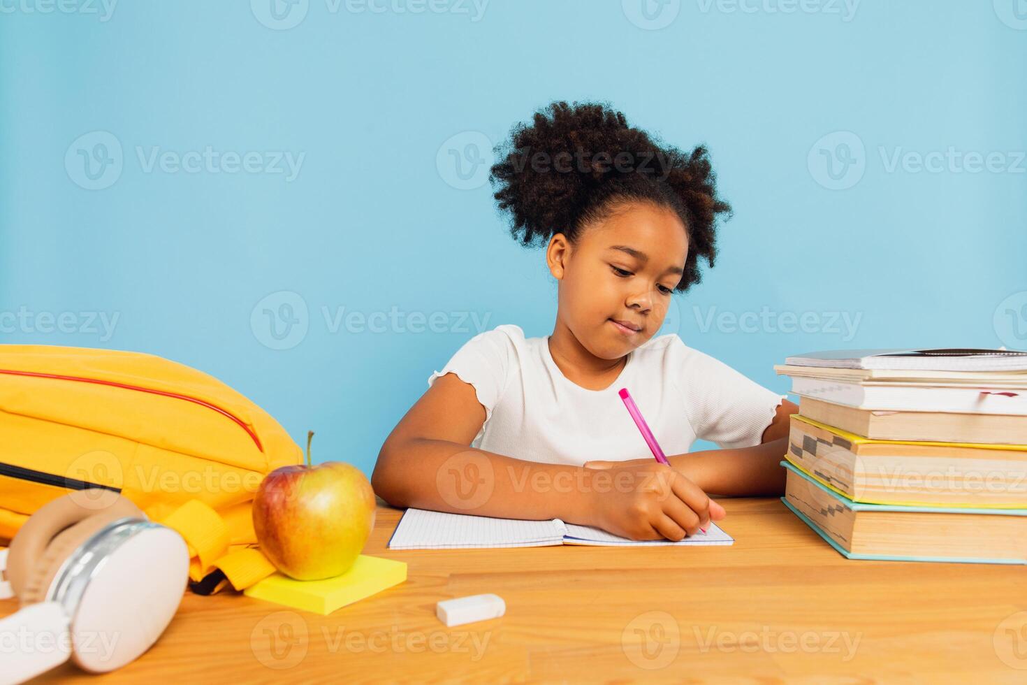 Happy African American schoolgirl doing homework at desk in class on blue background. Back to school concept. photo