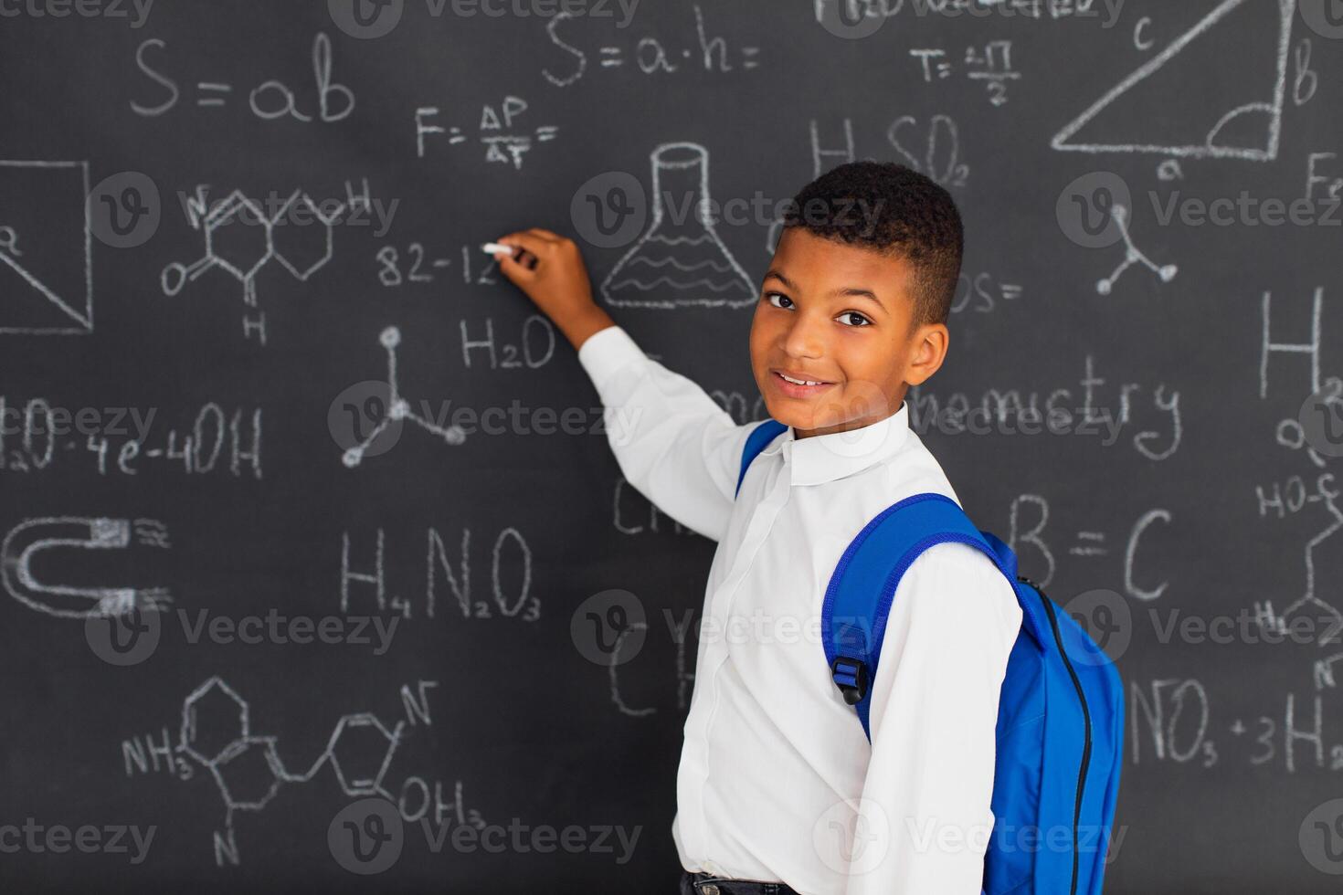 Happy mixed race schoolboy solving problems near the blackboard at school, back to school concept. photo