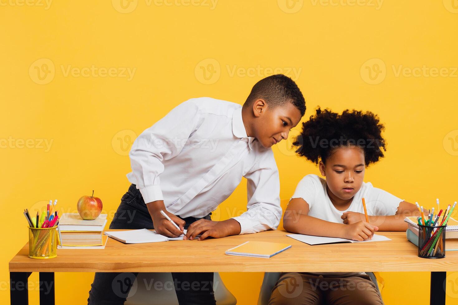 Happy african american schoolgirl and mixed race schoolboy sitting together at desk and studying on yellow background. Back to school concept. photo