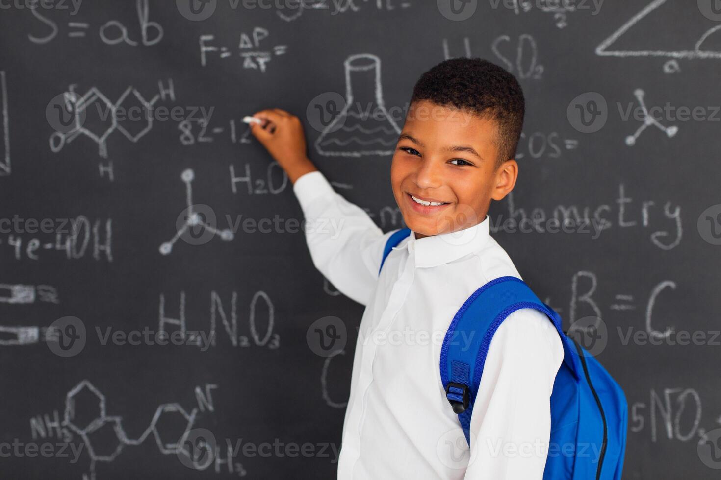 Happy mixed race schoolboy solving problems near the blackboard at school, back to school concept. photo