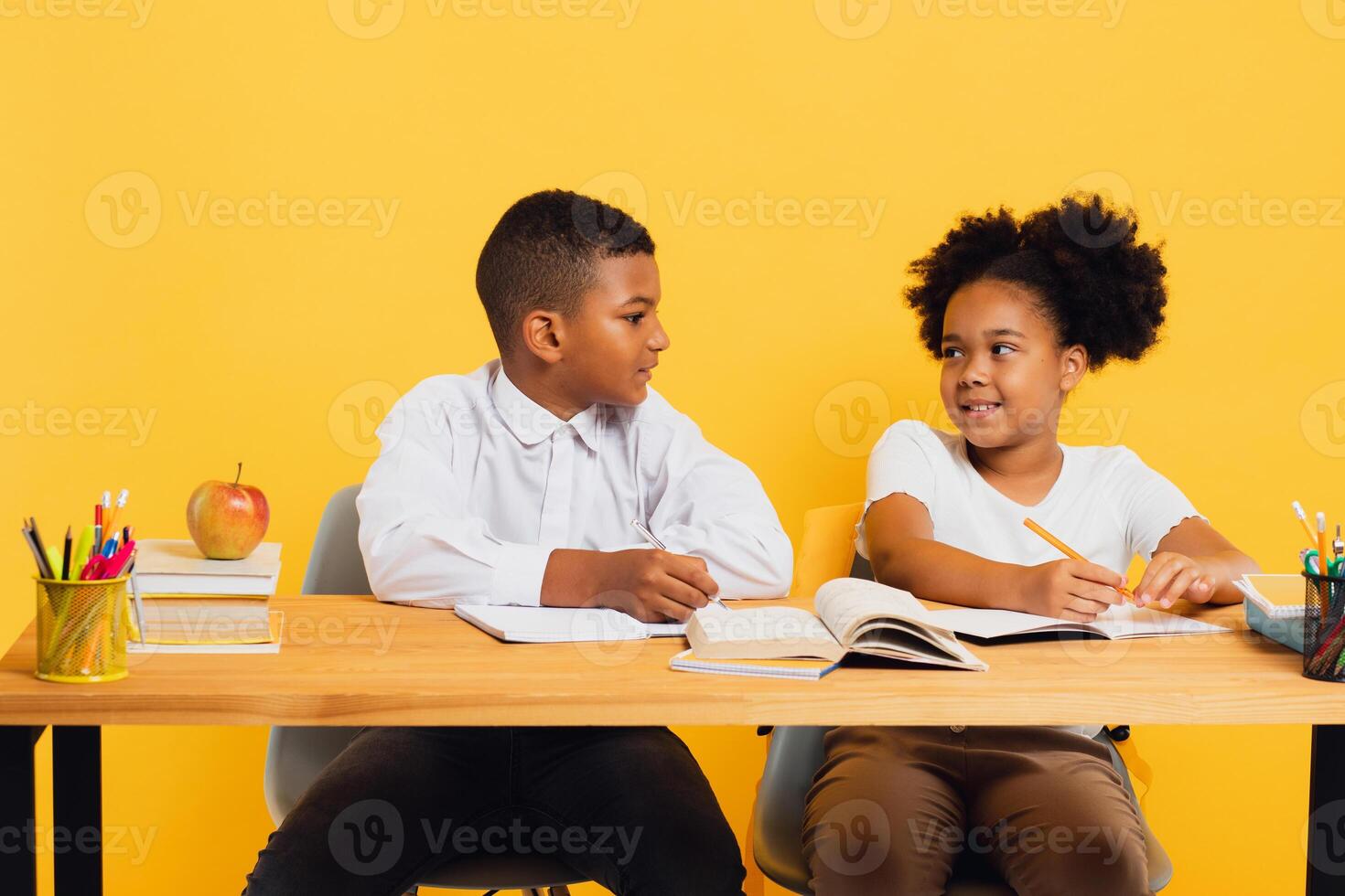 Happy african american schoolgirl and schoolboy sitting together at desk and studying on yellow background. Back to school concept. photo