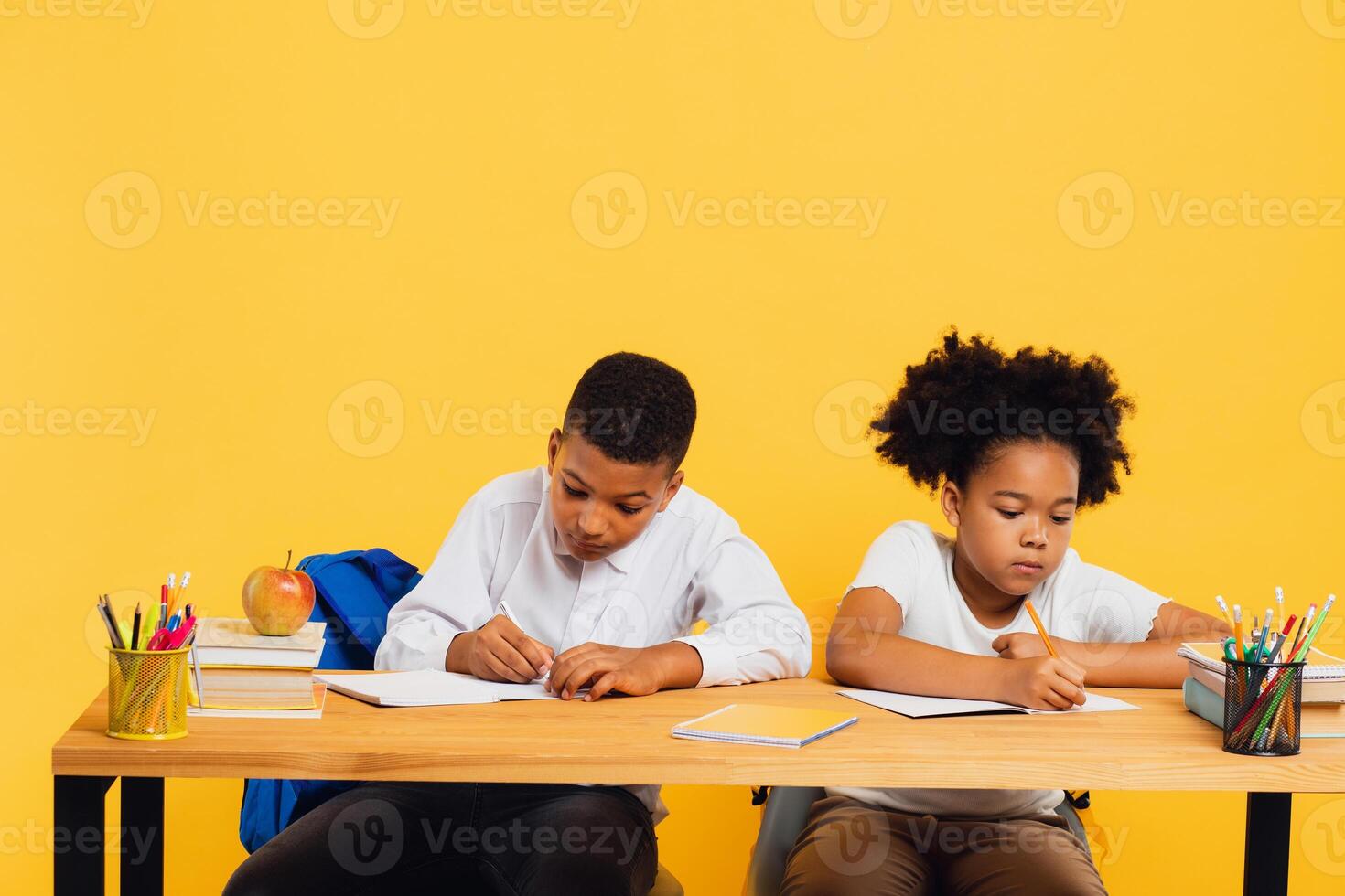 Happy african american schoolgirl and mixed race schoolboy sitting together at desk and studying on yellow background. Back to school concept. photo