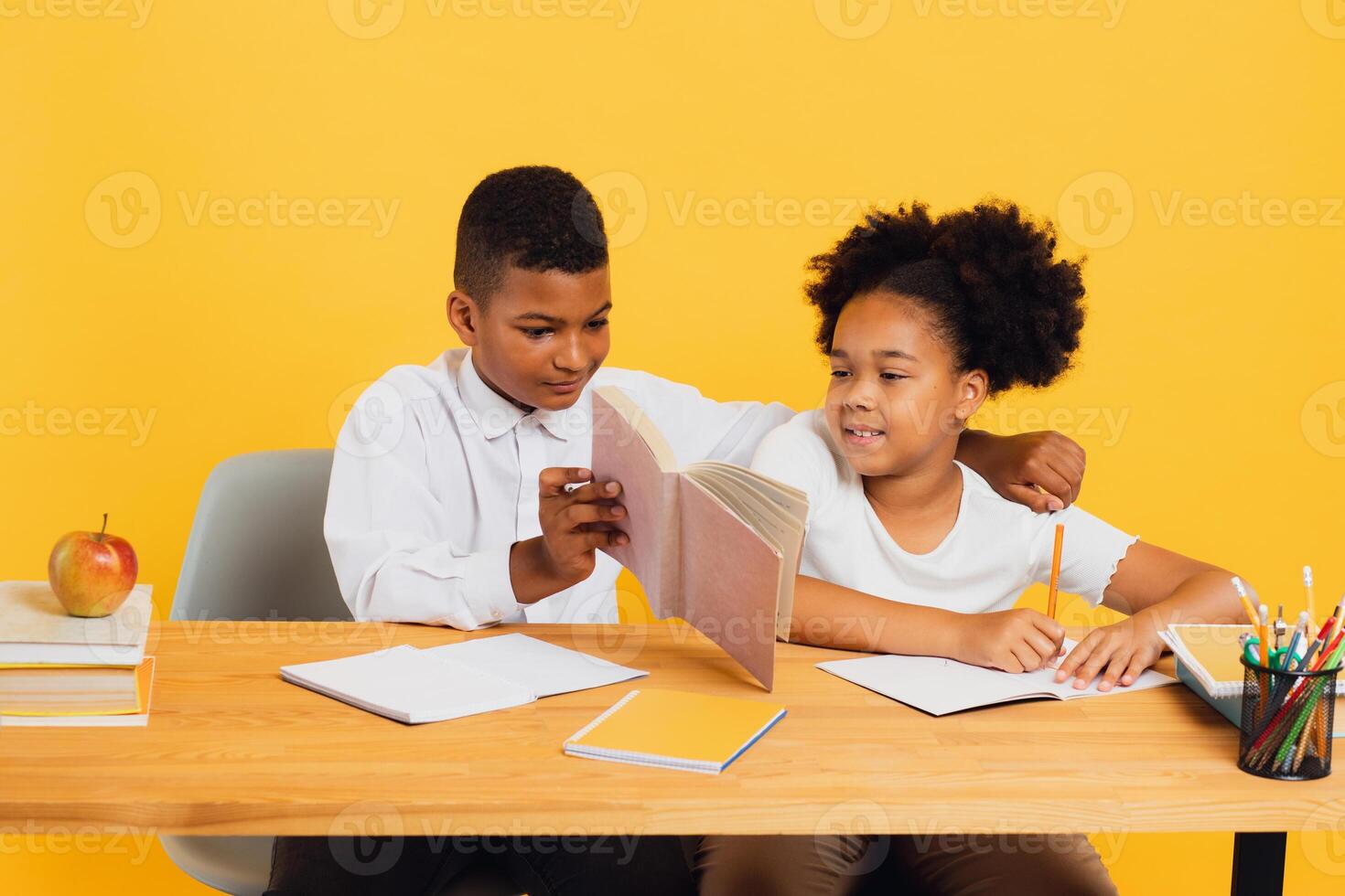 Happy african american schoolgirl and schoolboy sitting together at desk and studying on yellow background. Back to school concept. photo