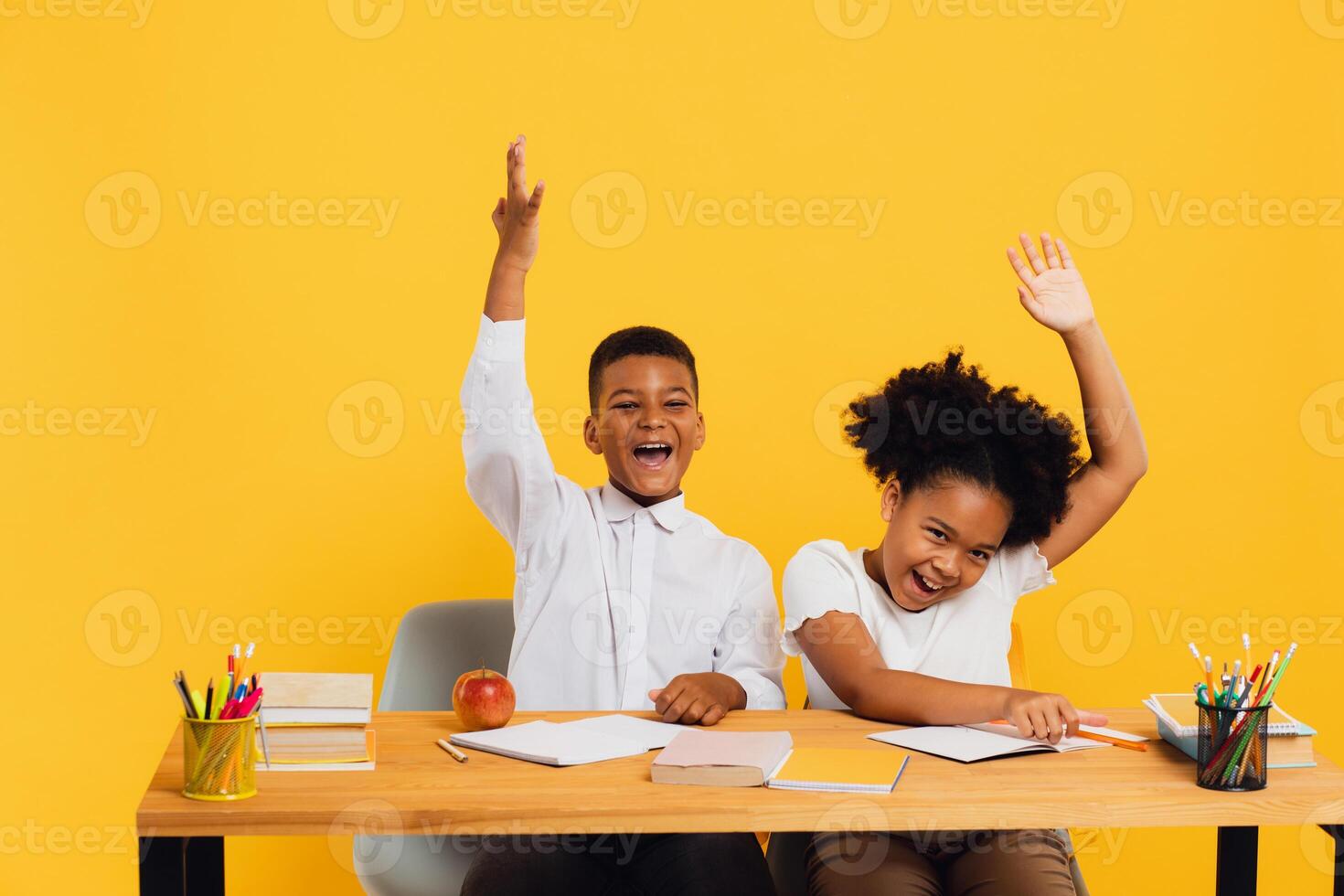 Happy african american female schoolgirl and schoolboy sitting together at desk and stretching arms up on yellow background. Back to school concept. photo