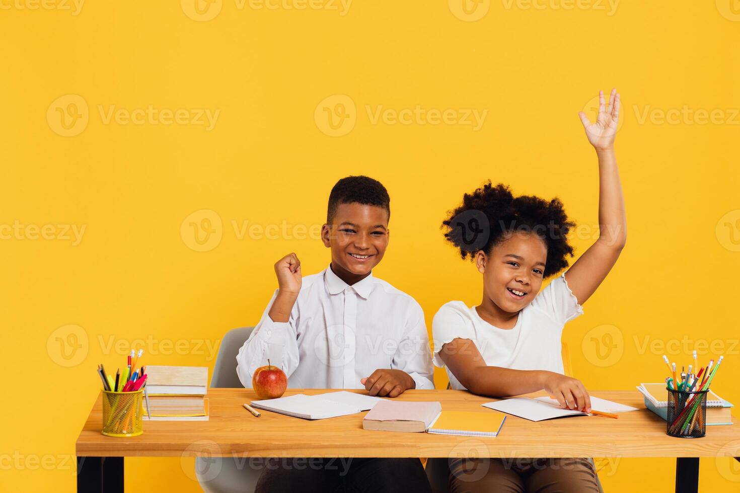 Happy african american female schoolgirl and schoolboy sitting together at desk and stretching arms up on yellow background. Back to school concept. photo