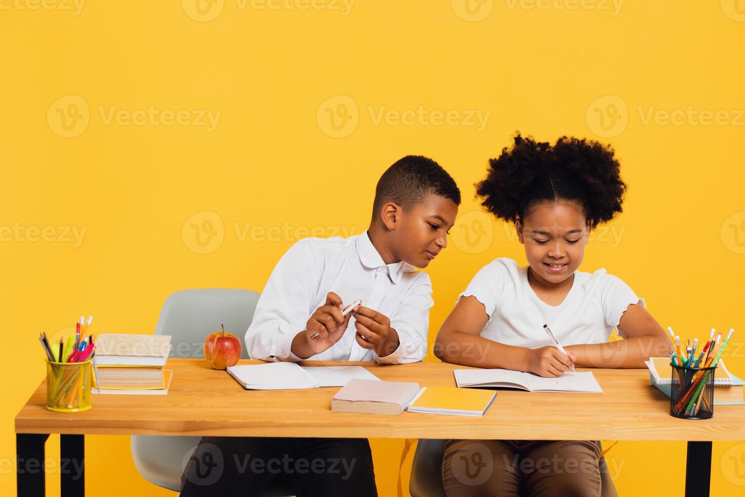 Happy african american schoolgirl and schoolboy sitting together at desk and studying on yellow background. Back to school concept. photo