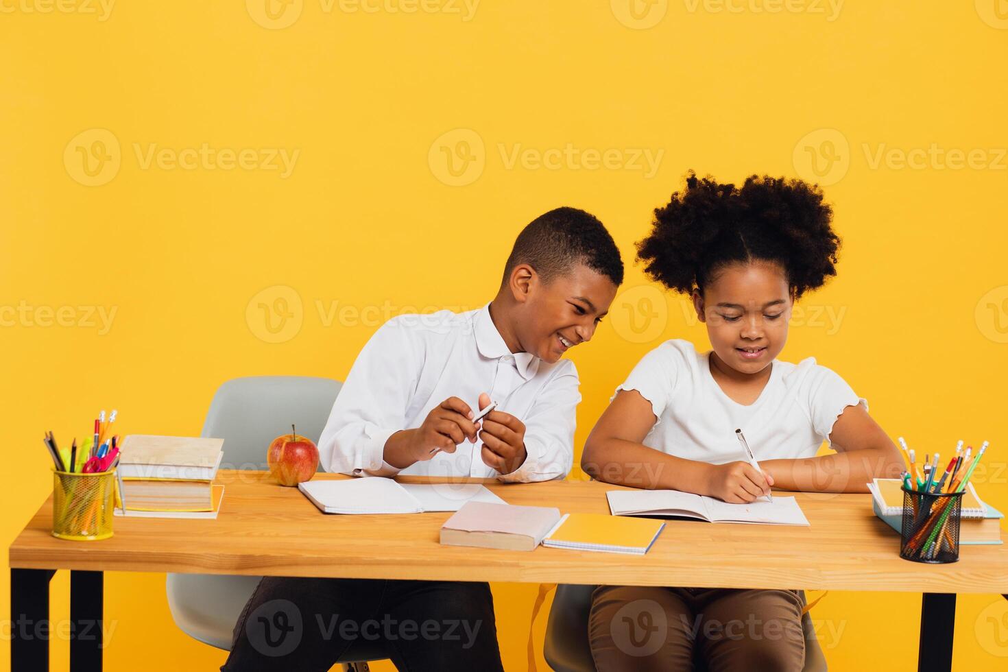 Happy african american schoolgirl and schoolboy sitting together at desk and studying on yellow background. Back to school concept. photo