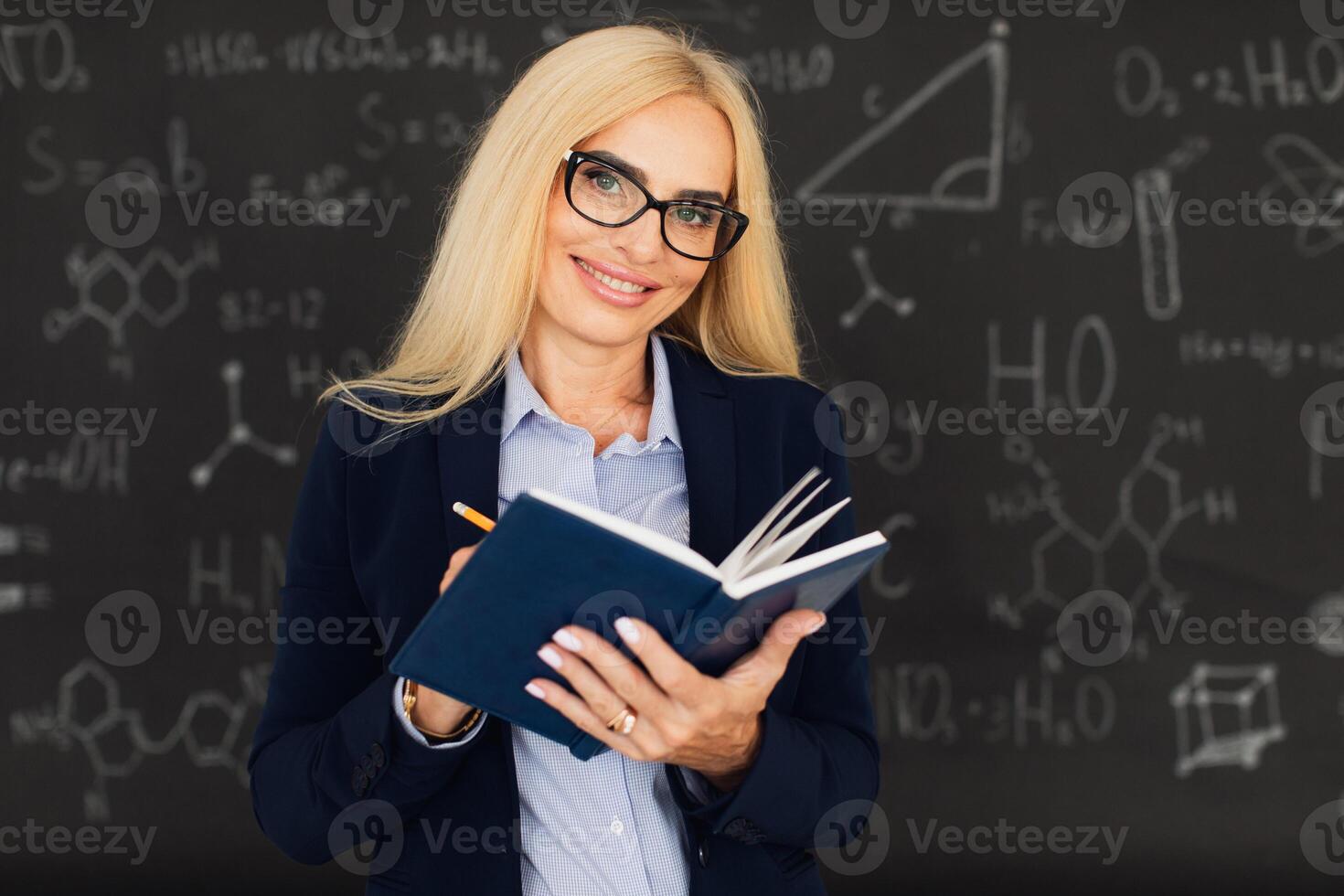 Portrait of happy blonde teacher woman standing near the blackboard in the classroom. photo