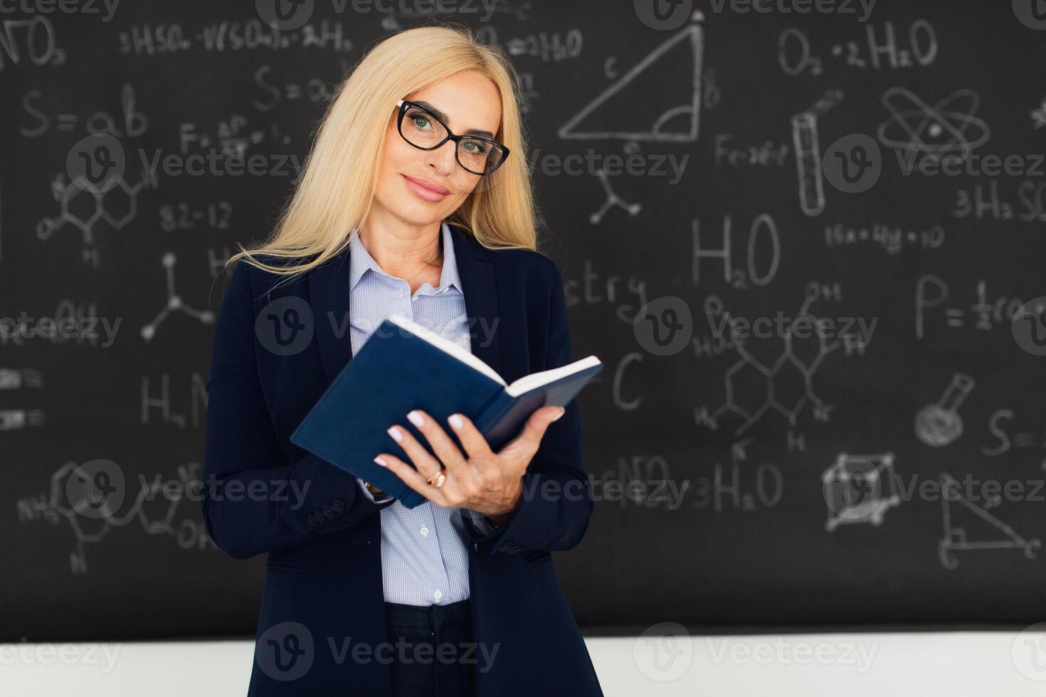 Portrait of happy blonde teacher woman standing near the blackboard in the classroom. photo