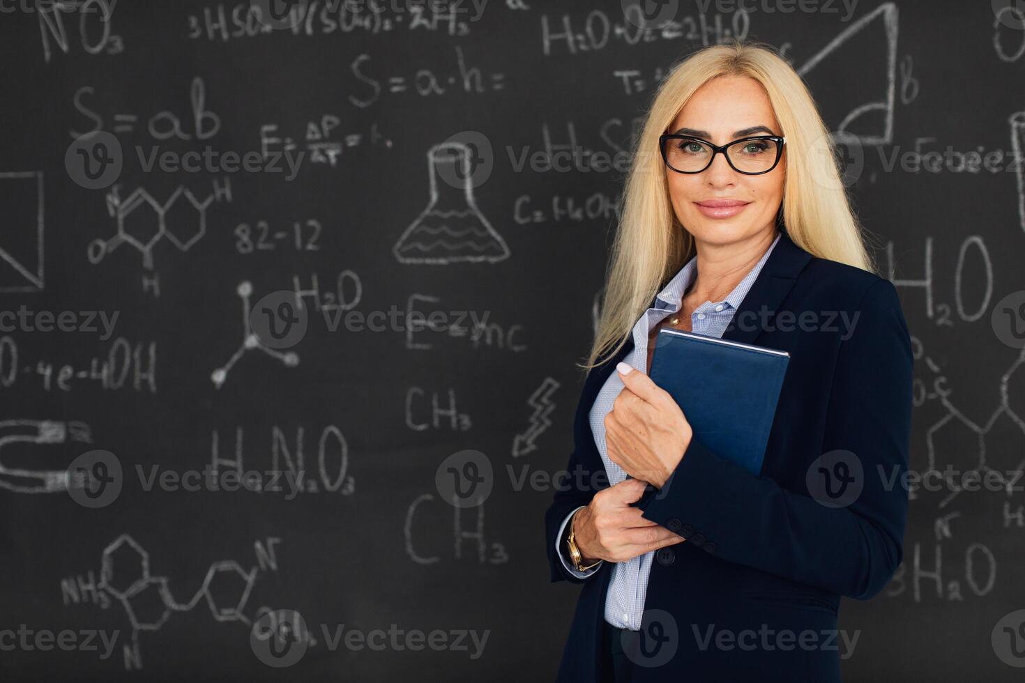 Portrait of happy blonde teacher woman standing near the blackboard in the classroom. photo