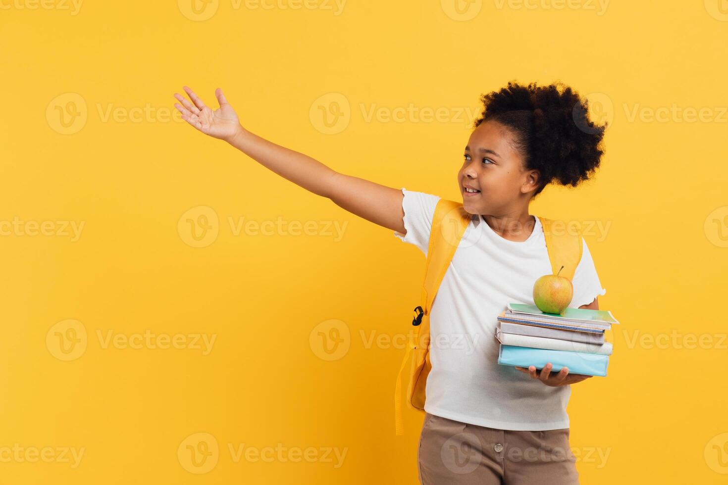 African american schoolgirl holding notebooks and books and showing something on yellow background, copy space. photo