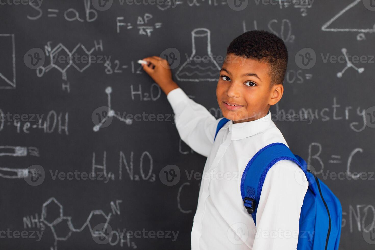 Happy african schoolboy solving problems near the blackboard at school, back to school concept photo