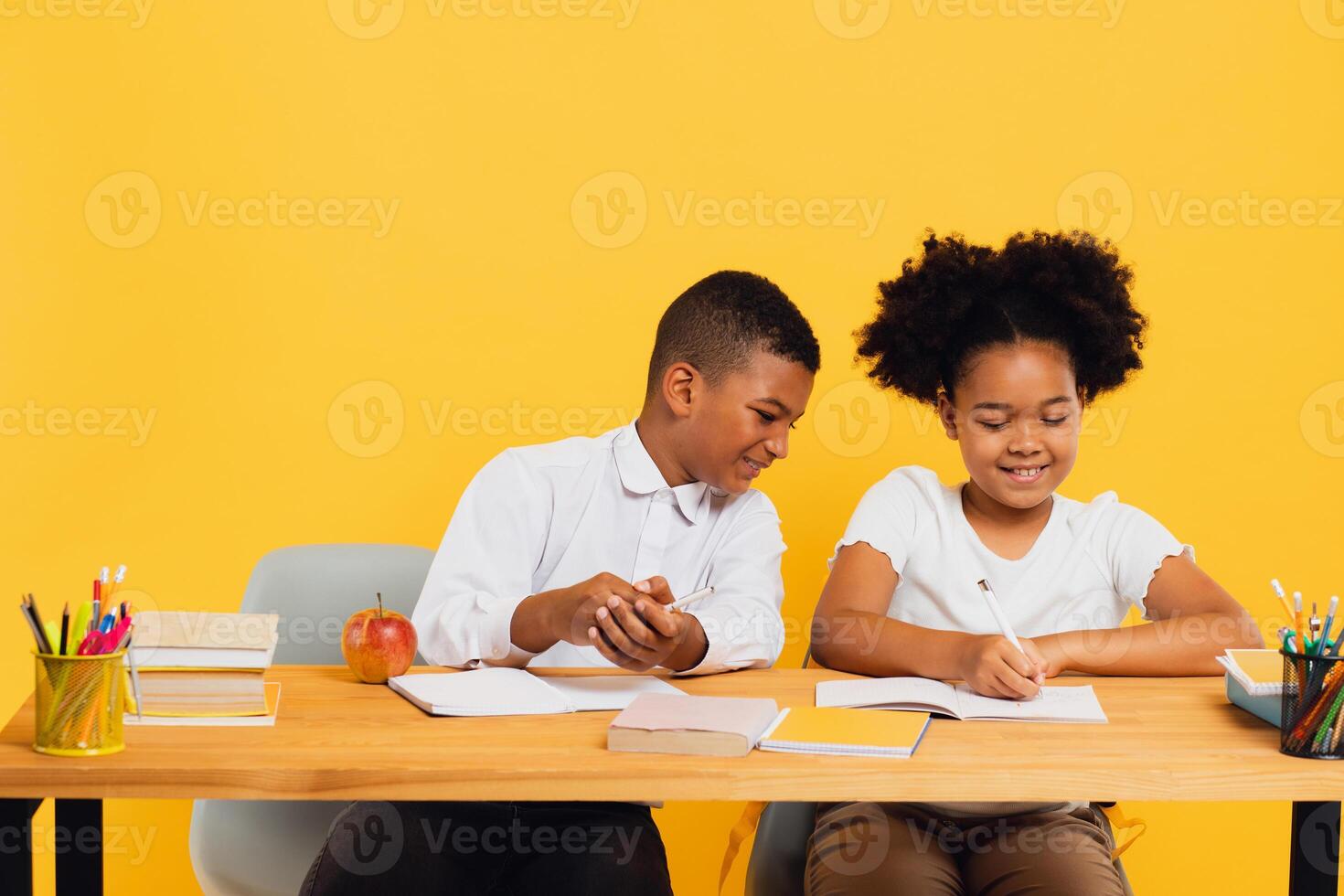 Happy african american schoolgirl and schoolboy sitting together at desk and studying on yellow background. Back to school concept photo