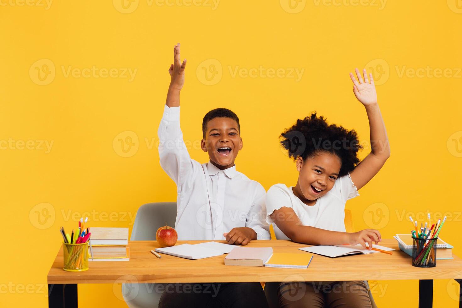 Happy african american female schoolgirl and schoolboy sitting together at desk and stretching arms up on yellow background. Back to school concept photo