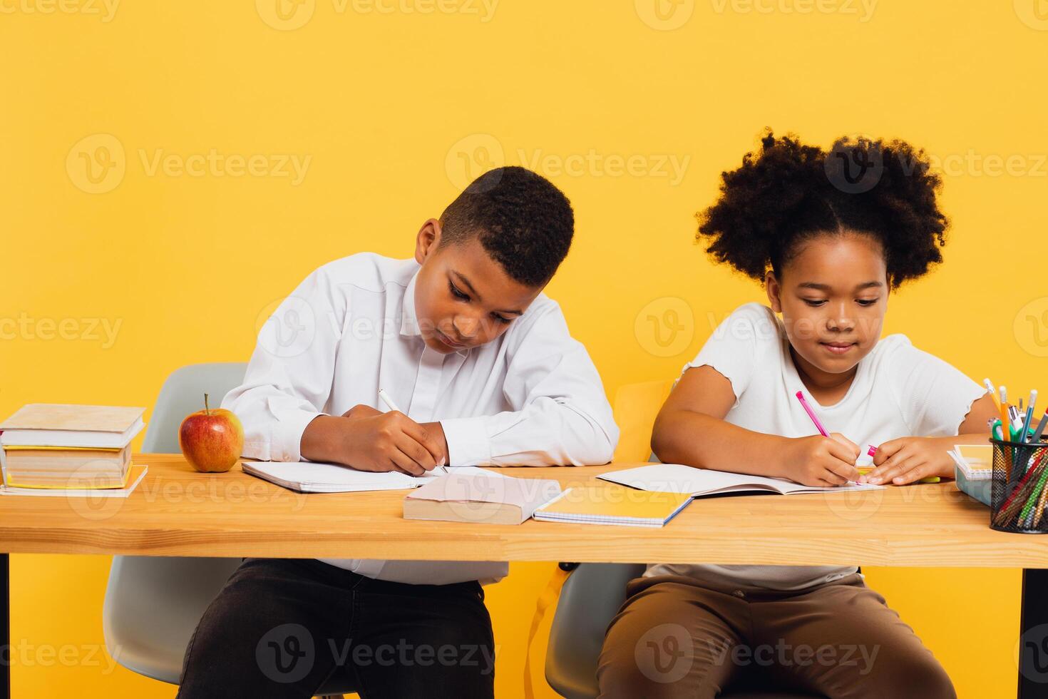 Happy african american schoolgirl and schoolboy sitting together at desk and studying on yellow background. Back to school concept photo