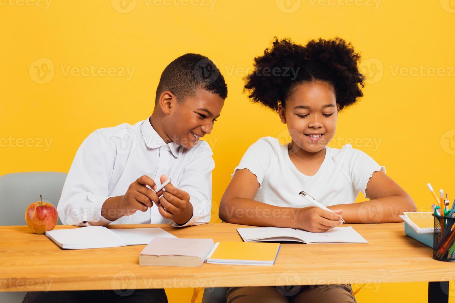 Happy african american schoolgirl and schoolboy sitting together at desk and studying on yellow background. Back to school concept photo