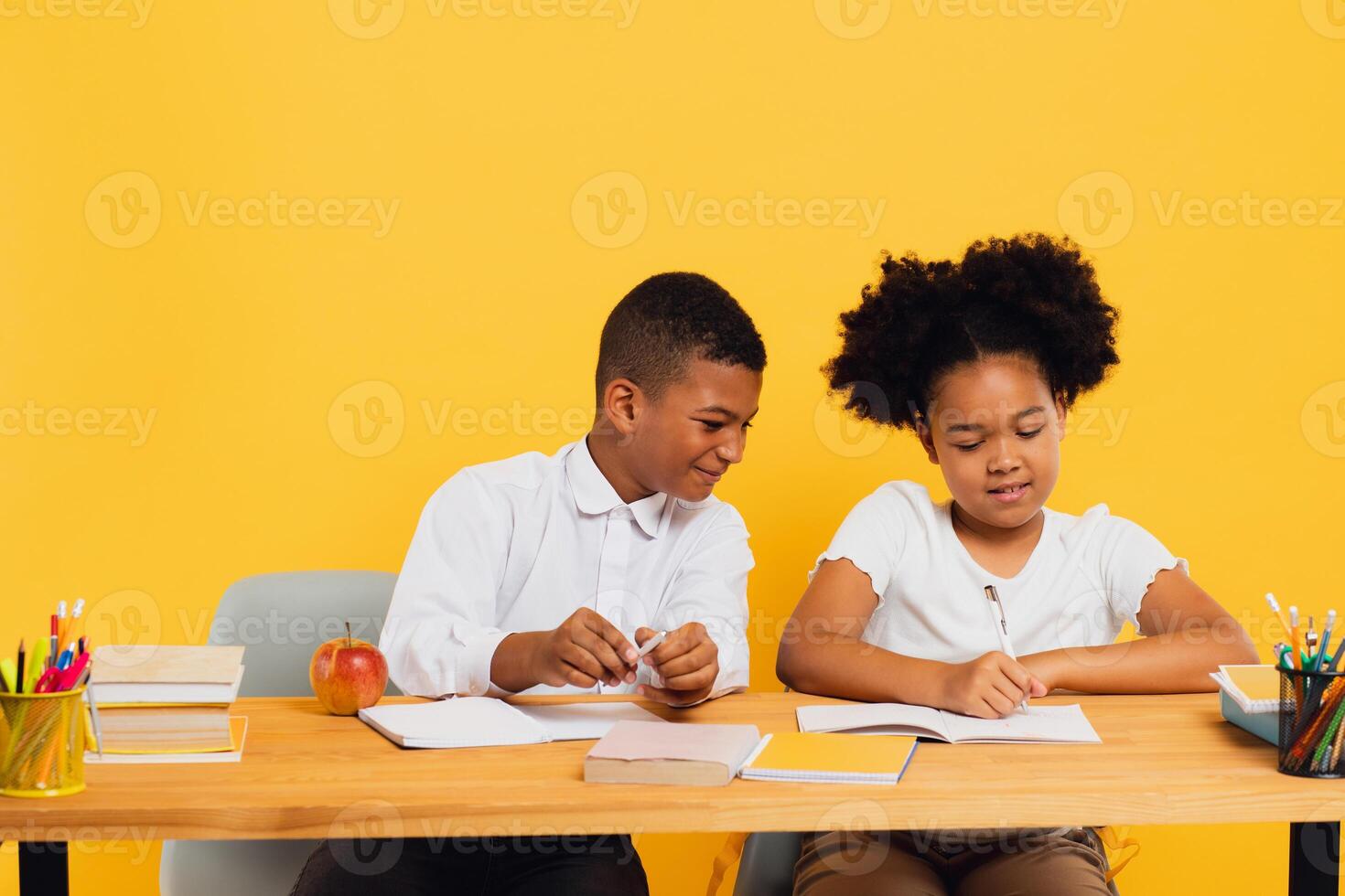 Happy african american schoolgirl and schoolboy sitting together at desk and studying on yellow background. Back to school concept photo