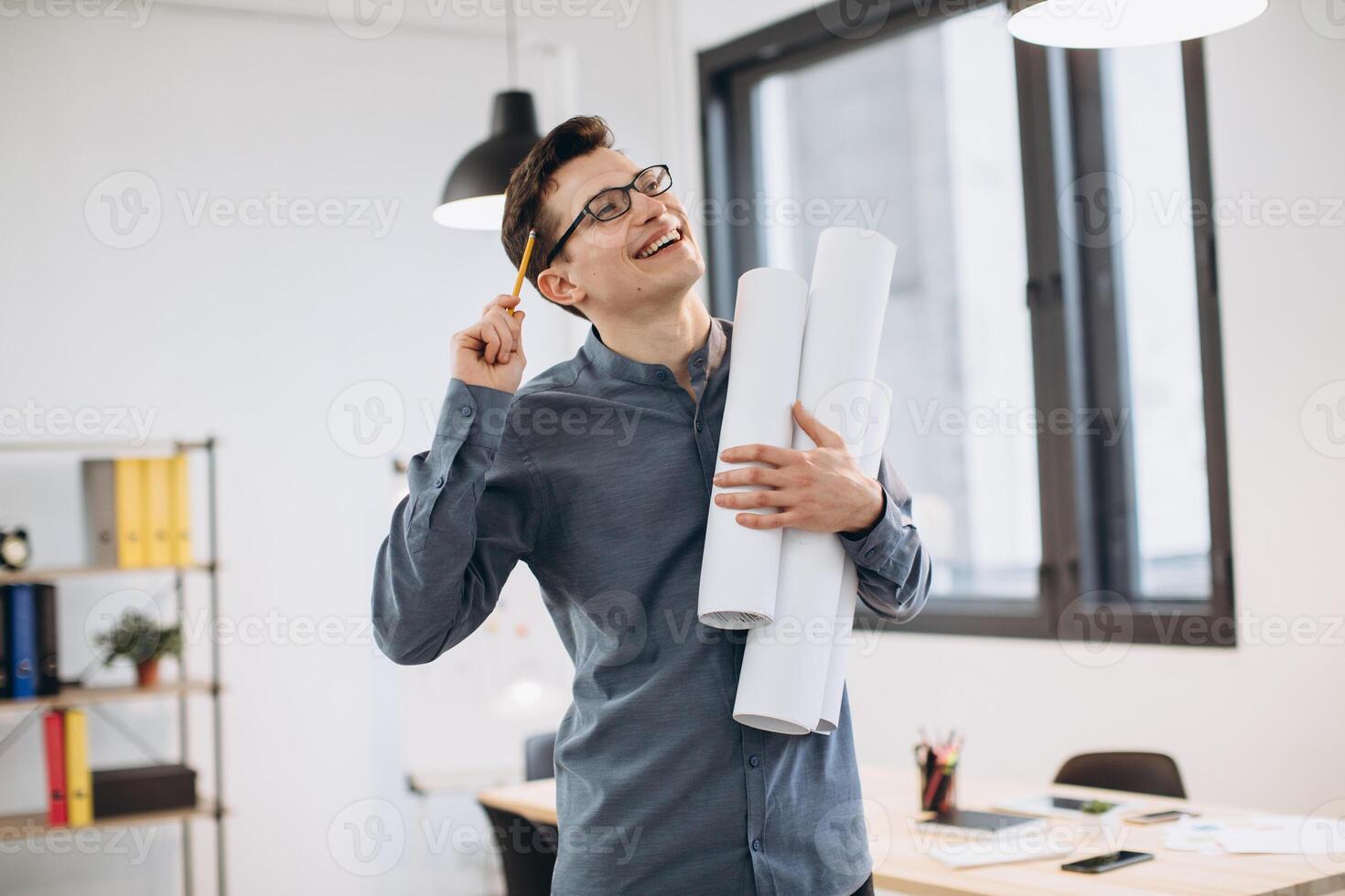 Attractive young man architect in glasses standing isolated over loft office background, carrying drawings, holding house scheme photo