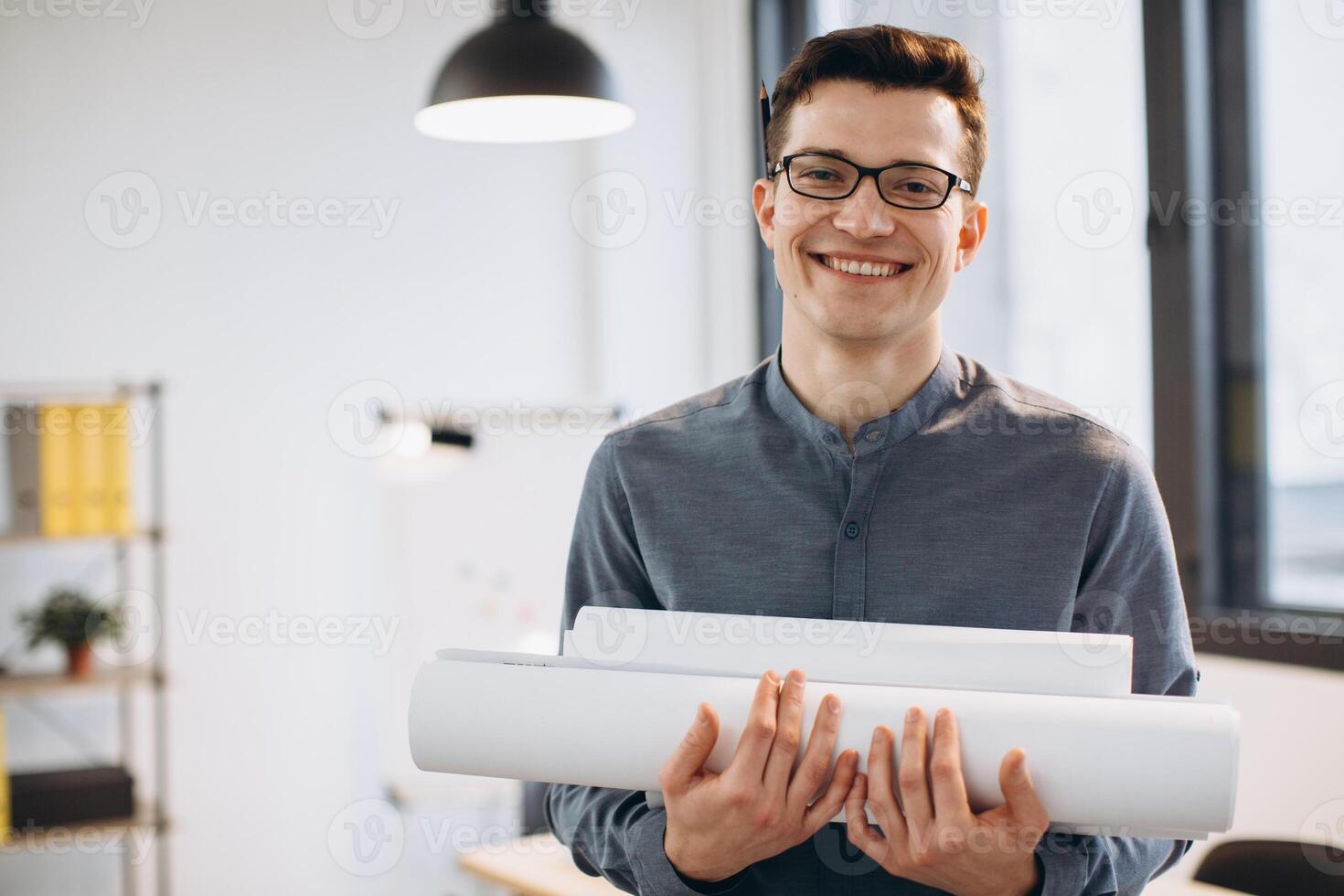 Attractive young man architect in glasses standing isolated over loft office background, carrying drawings, holding house scheme photo