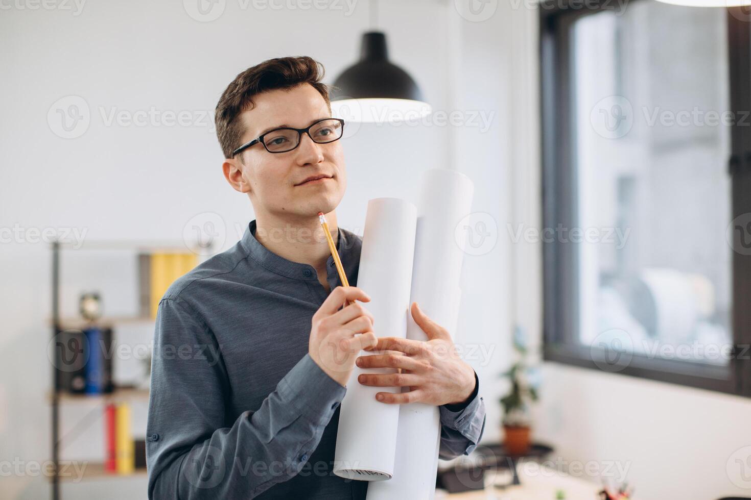 Attractive young man architect in glasses standing isolated over loft office background, carrying drawings, holding house scheme photo