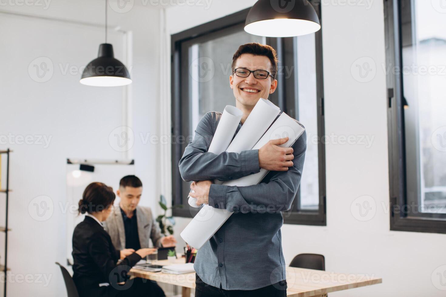 Attractive young man architect in glasses standing isolated over loft office background, carrying drawings, holding house scheme photo