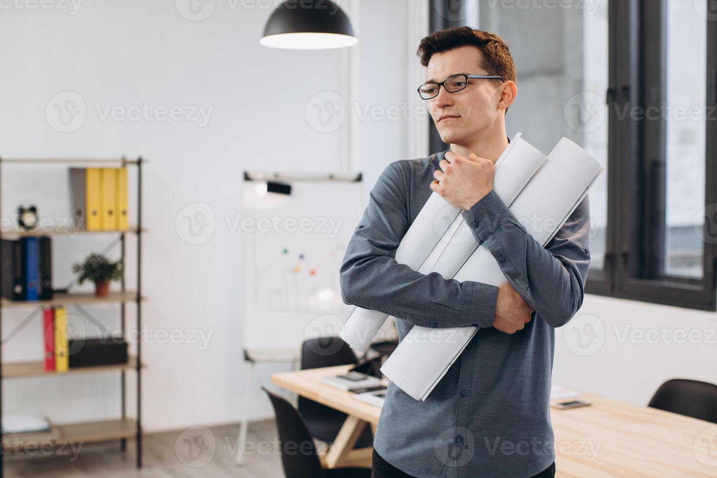Attractive young man architect in glasses standing isolated over loft office background, carrying drawings, holding house scheme photo