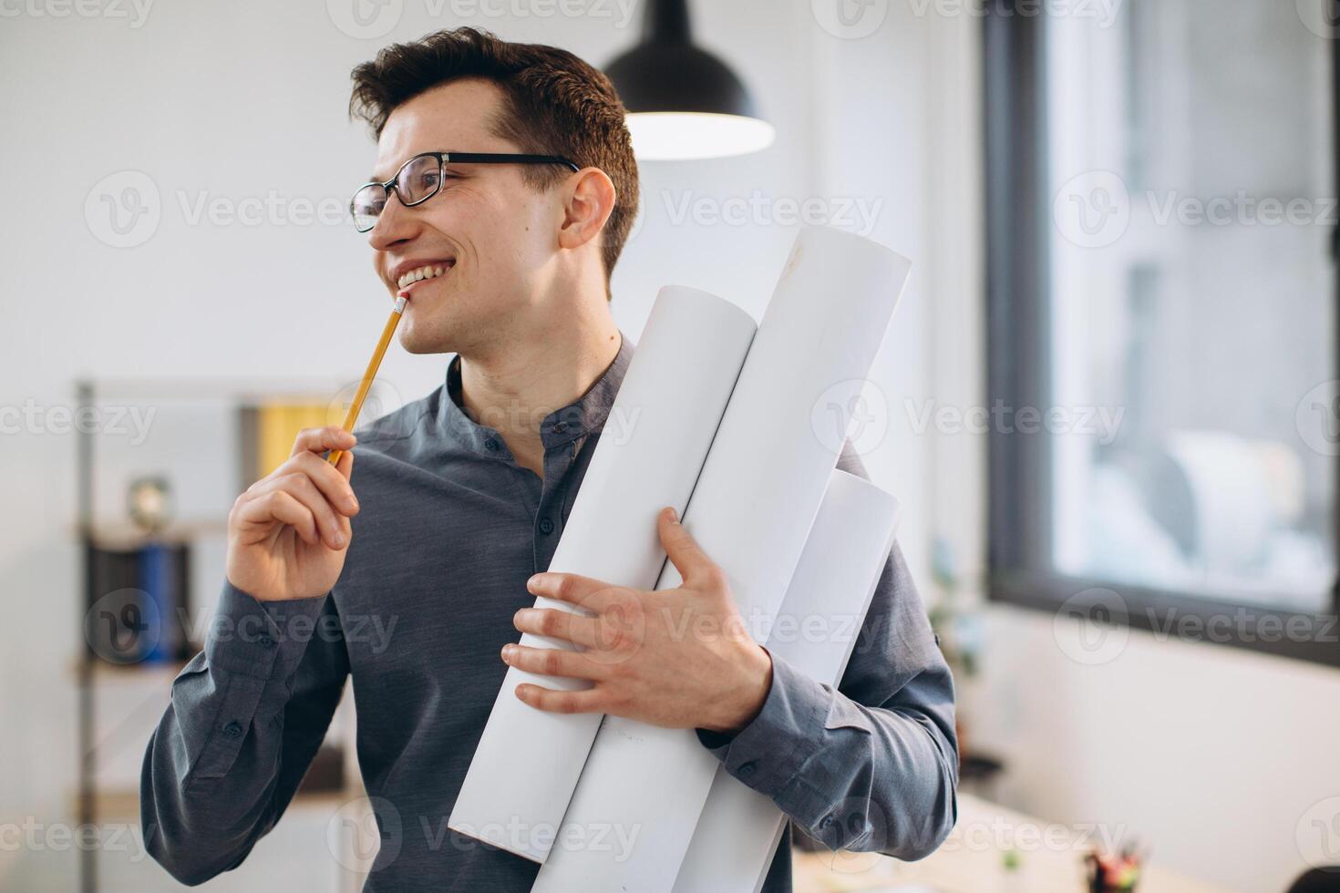Attractive young man architect in glasses standing isolated over loft office background, carrying drawings, holding house scheme photo