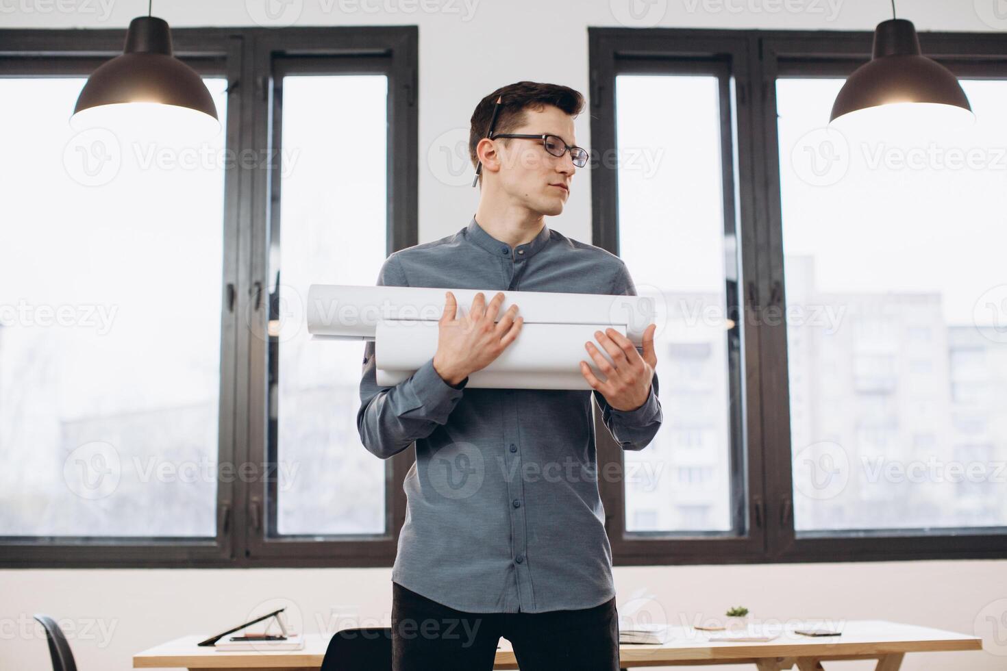 Attractive young man architect in glasses standing isolated over loft office background, carrying drawings, holding house scheme photo