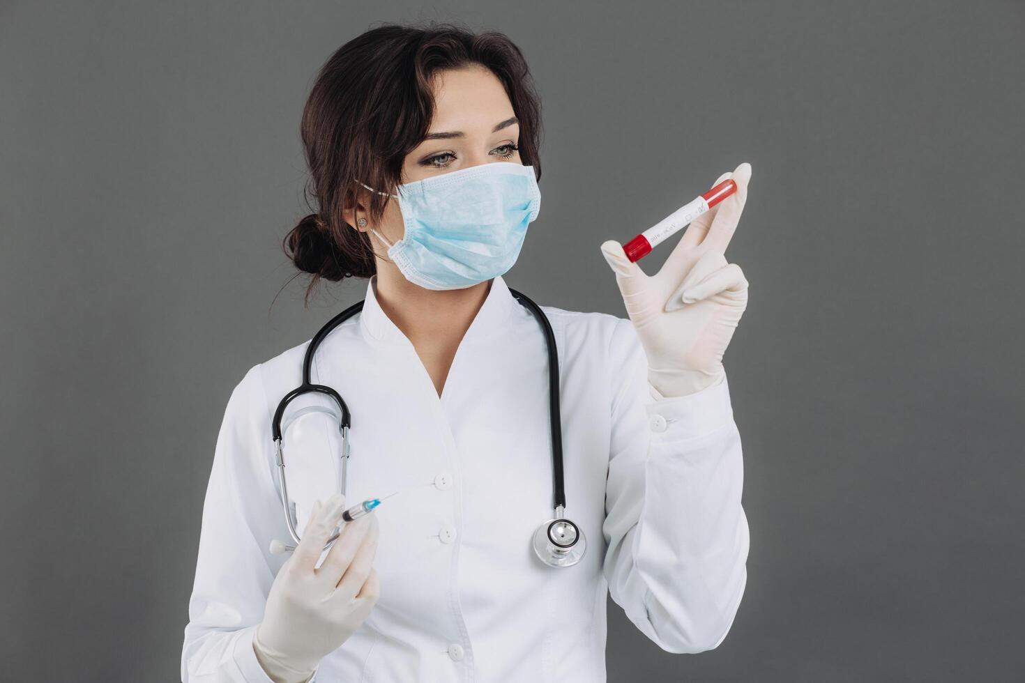 Female doctor with stethoscope in mask holding injection, antidote against coronavirus on gray background. photo