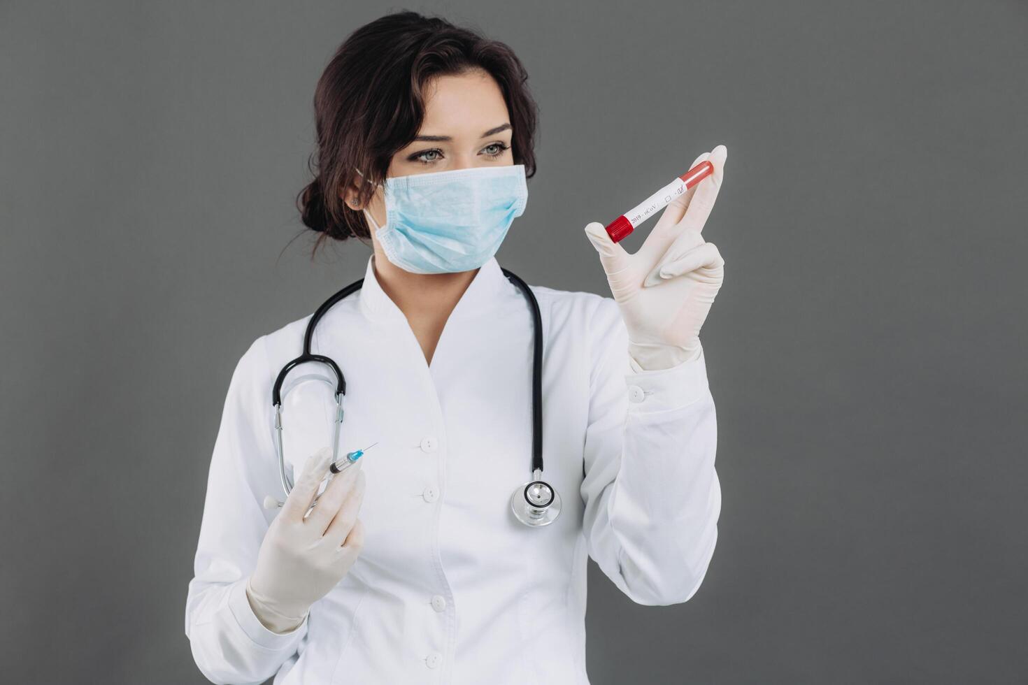 Female doctor with stethoscope in mask holding injection, antidote against coronavirus on gray background. photo