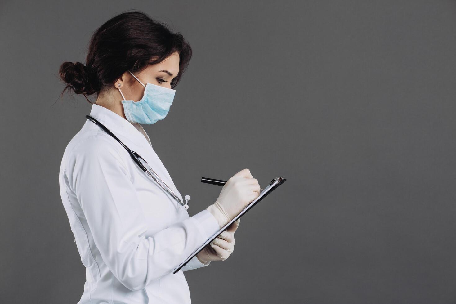 Woman doctor in mask and stethoscope holds folder and writes something in it related to coronavirus. Covid-19 concept photo