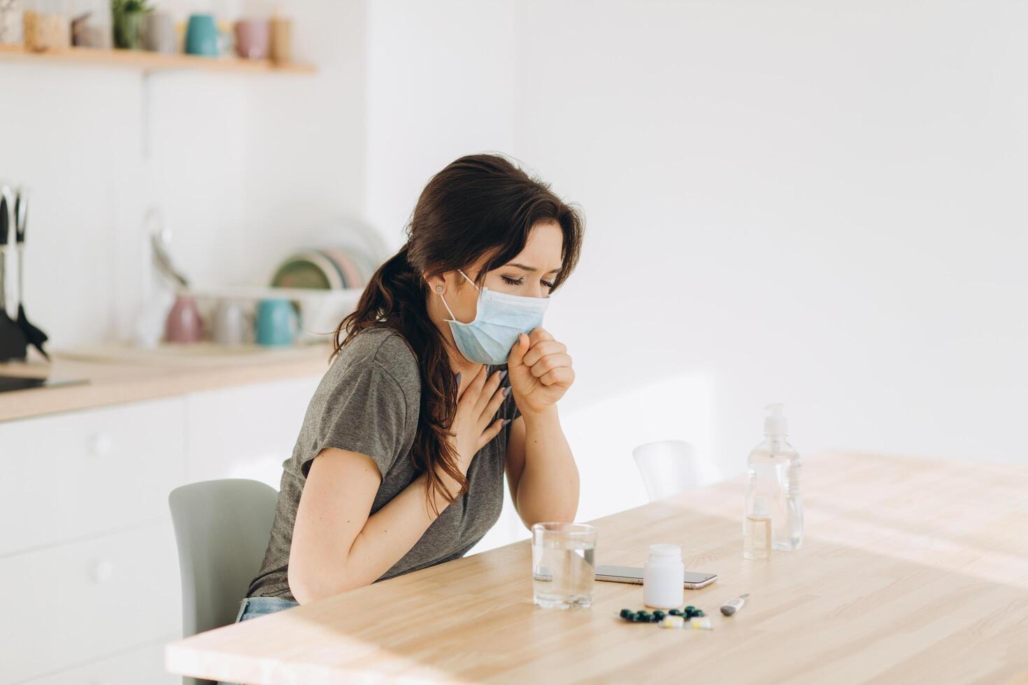 Woman in protective mask coughing in fist sick of coronavirus viral infection spreading coronavirus covering mouth and nose. Painful cough ill patient sitting in kitchen at home quarantine photo