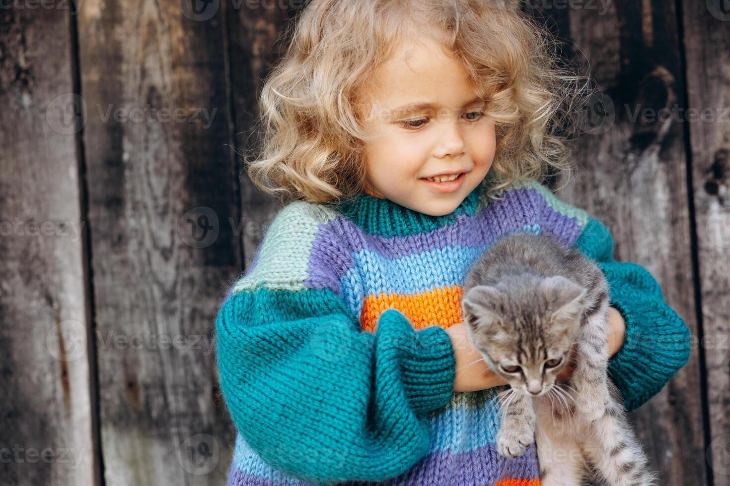 Portrait of a beautiful and happy curly girl in a knitted sweater playing with a kitten near a wooden wall photo