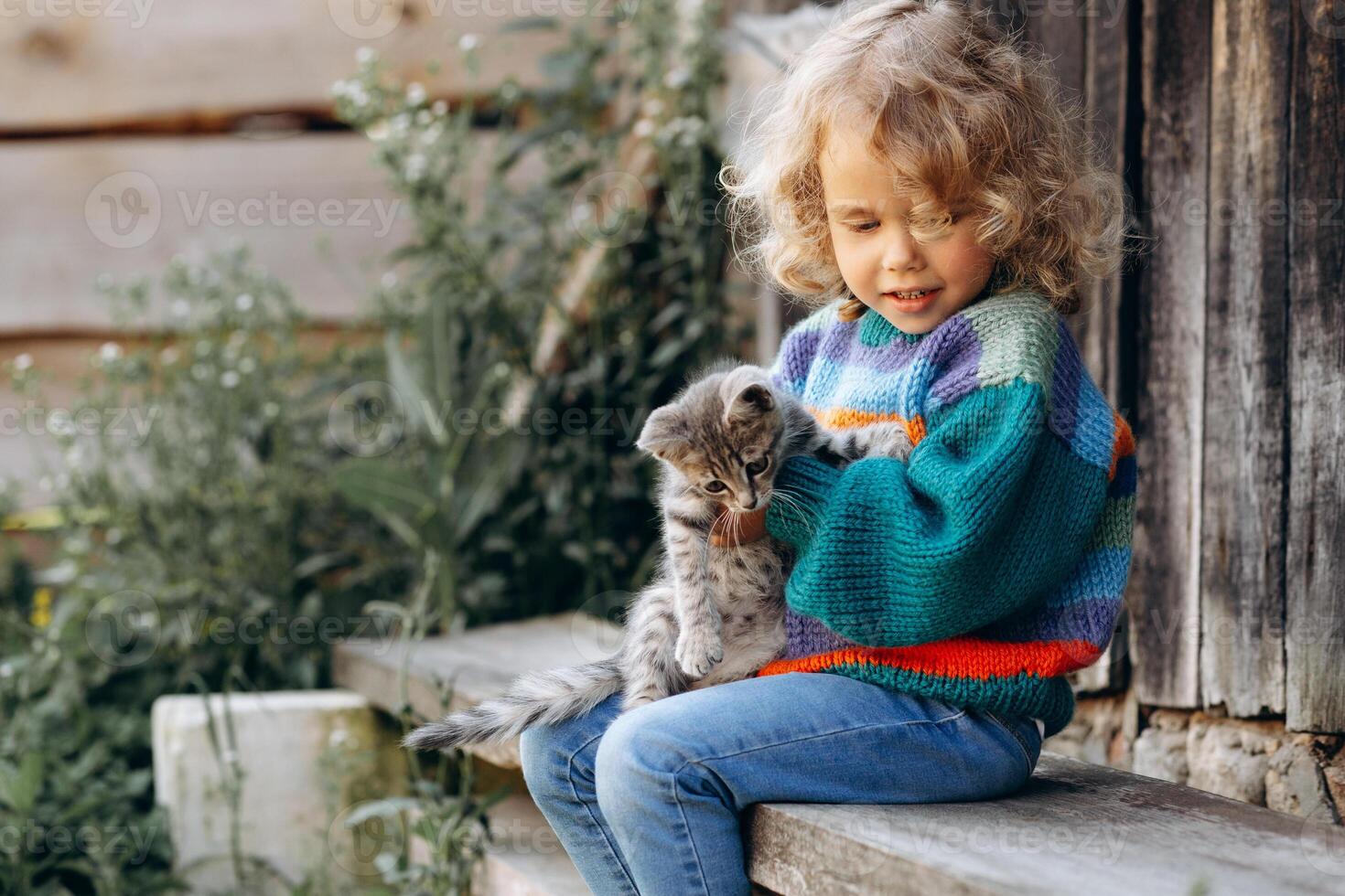 Portrait of a beautiful and happy curly girl in a knitted sweater playing with a kitten near a wooden wall photo