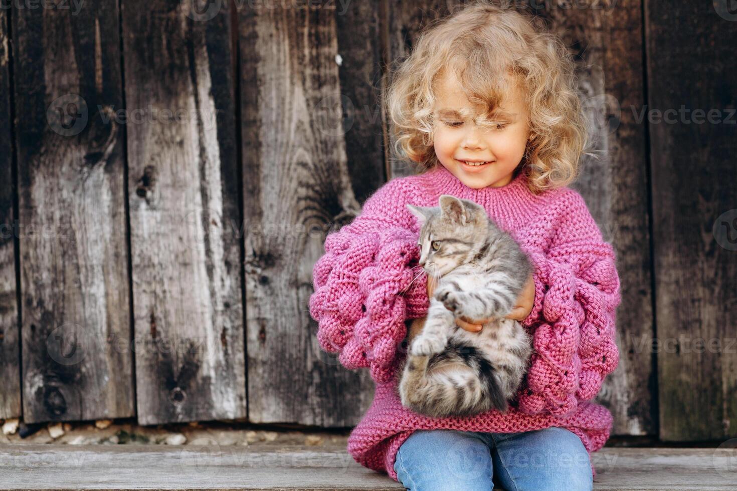 Portrait of a beautiful and happy curly girl in a knitted sweater playing with a kitten near a wooden wall photo