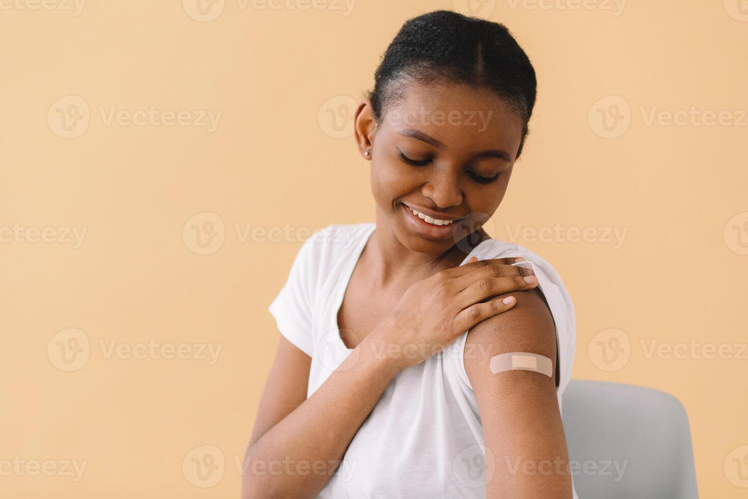 Young African American woman is happy about the first vaccination, wears band aid, raises sleeve and tells friends about the injection. Isolated over beige background. photo